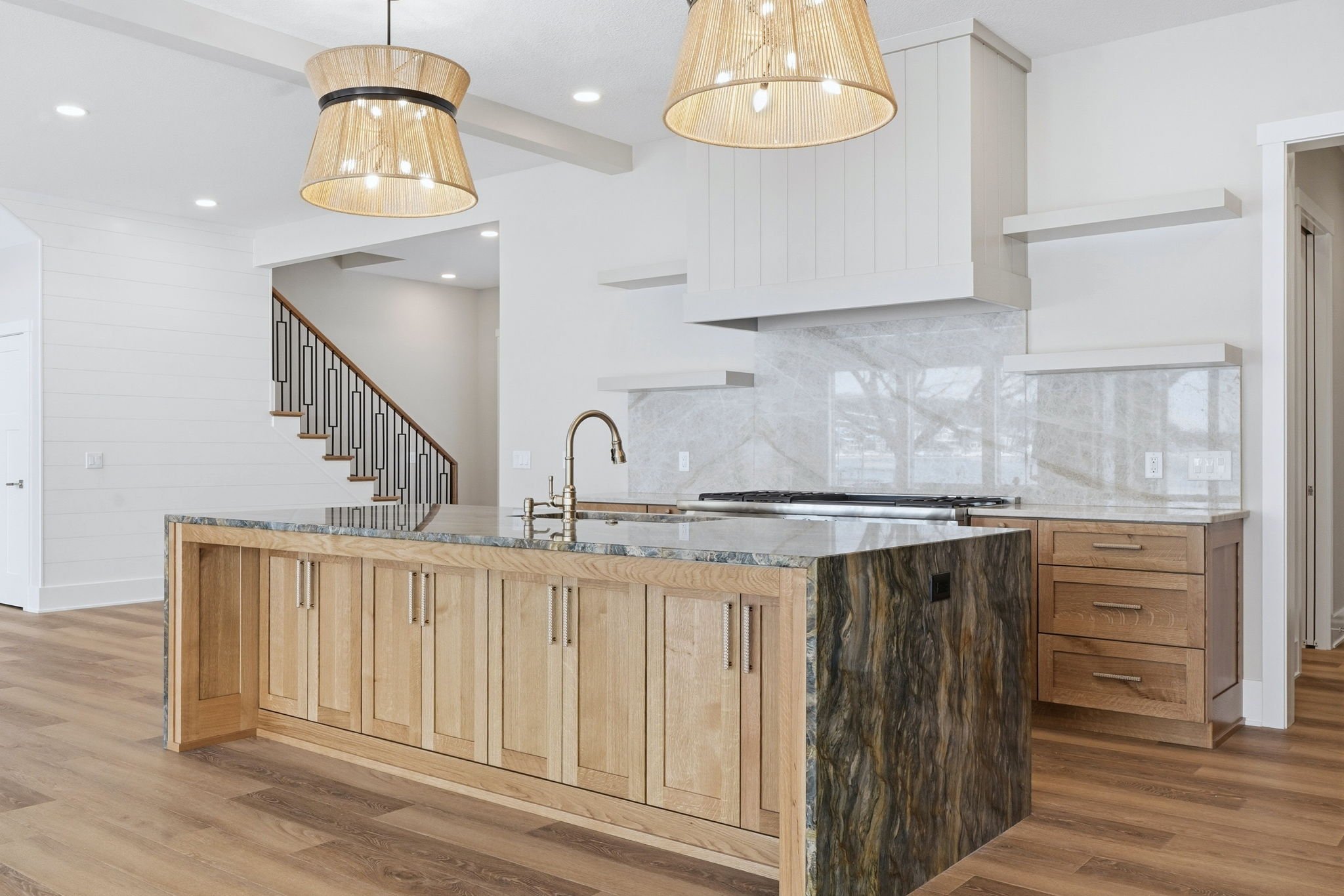 Modern kitchen with wooden island, light wood cabinetry, marble countertops, and pendant lighting, with a staircase visible in the background.