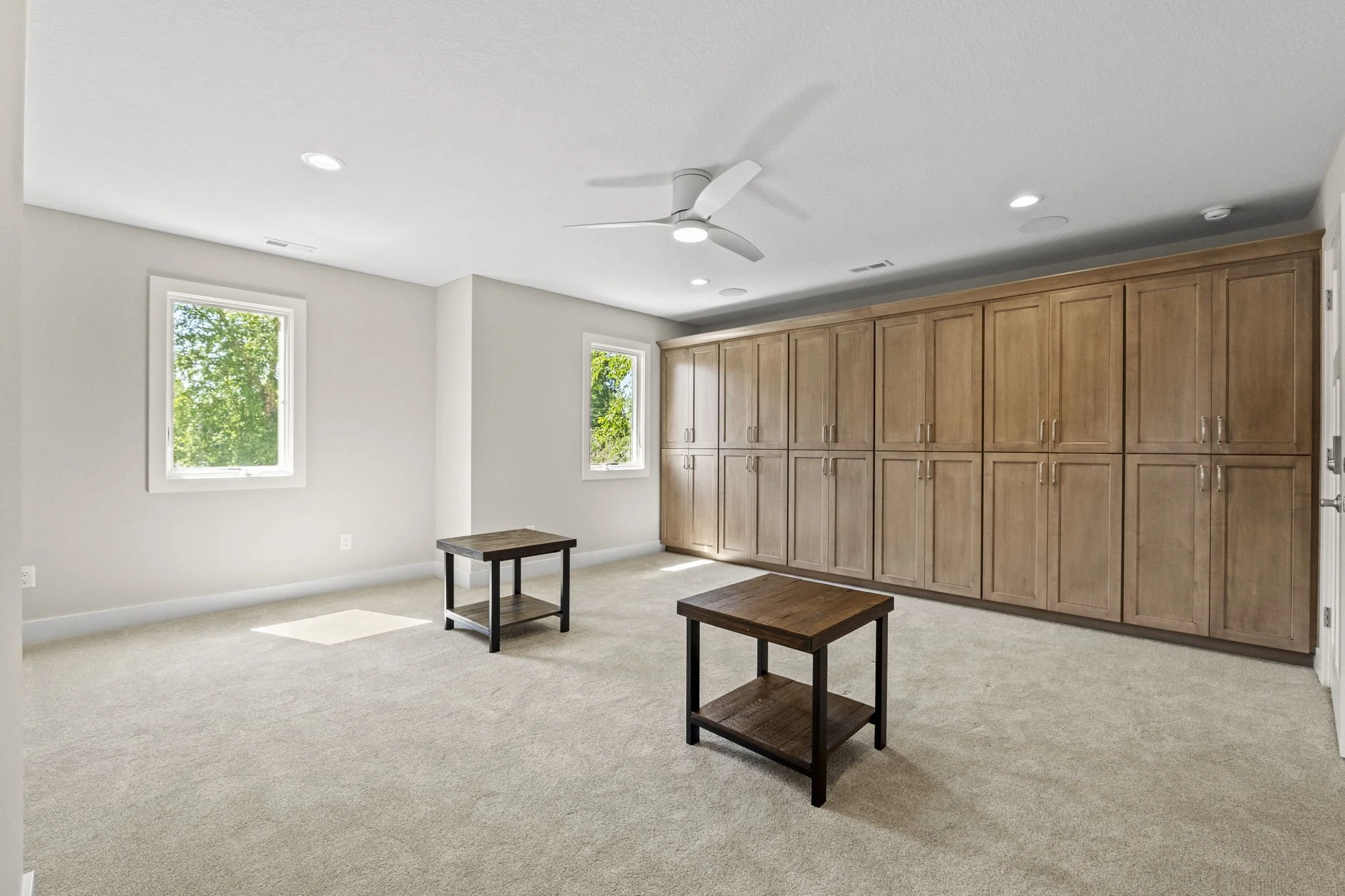 Empty bedroom with light-colored walls and carpet, two small wooden tables, large wooden closet on the right, two windows letting in natural light, ceiling fan, recessed lighting.
