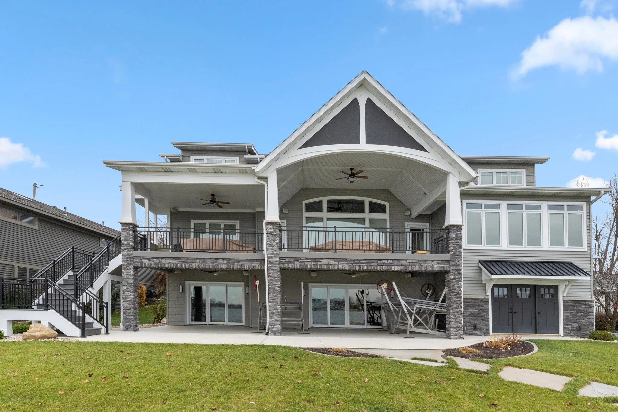 Large modern house with a spacious backyard patio, sliding glass doors, and a shaded upper balcony, with a manicured lawn and blue sky.