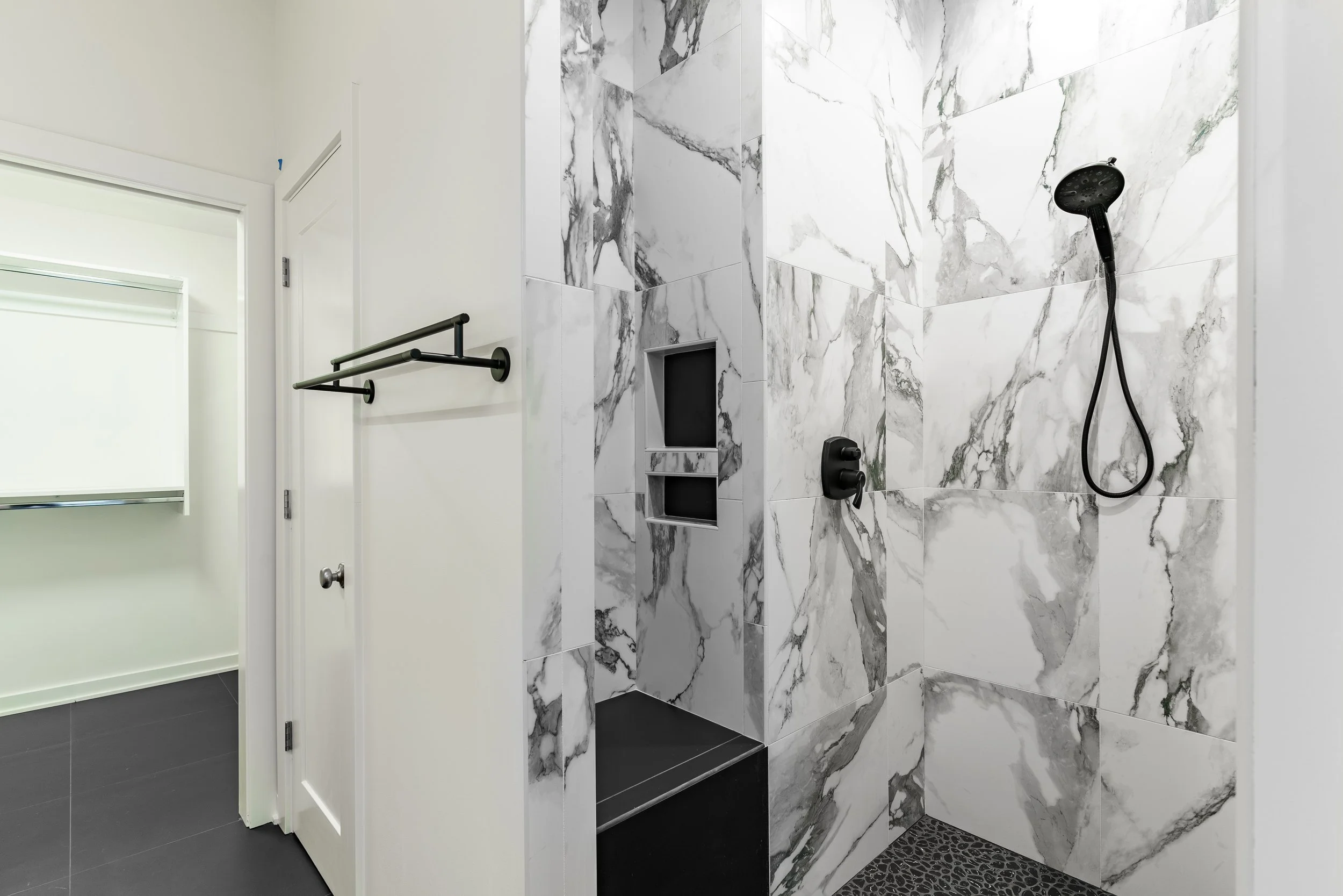 Bathroom shower area with white and gray marble tiles, black fixtures, a handheld showerhead, built-in soap shelves, and a black pebble floor.