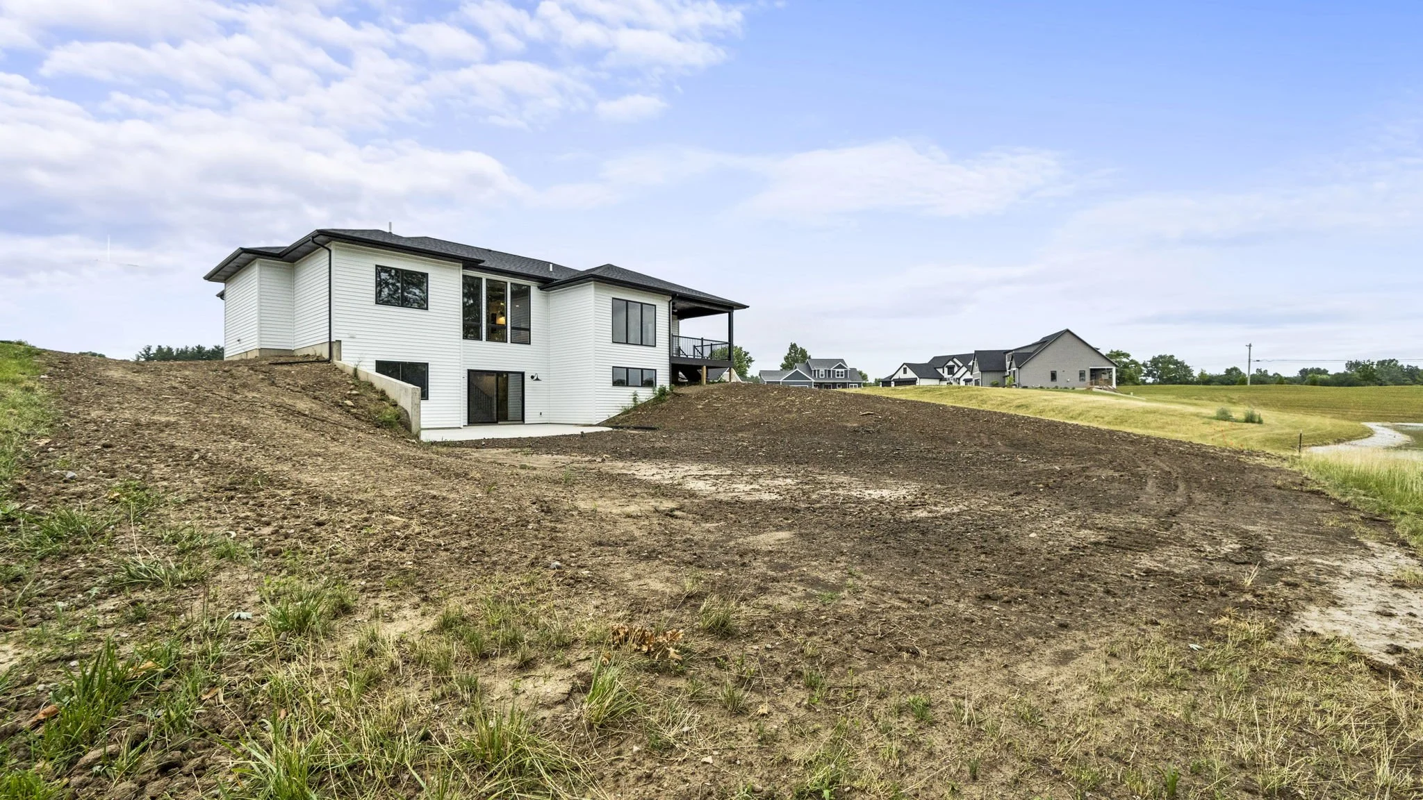 A modern white two-story house with large windows and a porch, situated on a sloped land with a dirt yard and surrounding grass, under a partly cloudy sky.