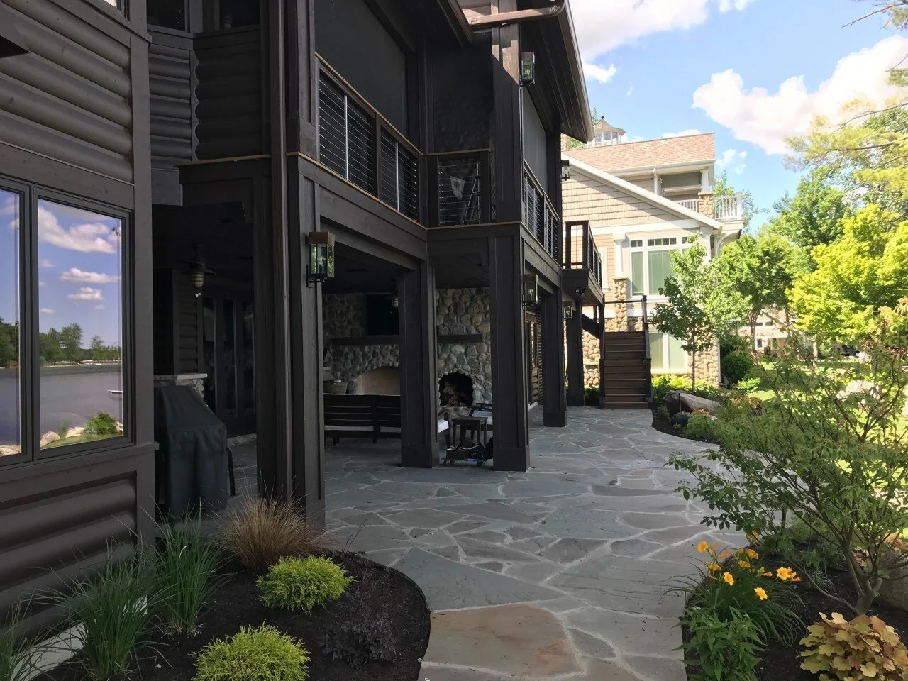 View of the backyard patio area with a stone pathway, outdoor fireplace, and a two-story house with a balcony and stairs, surrounded by greenery and trees.