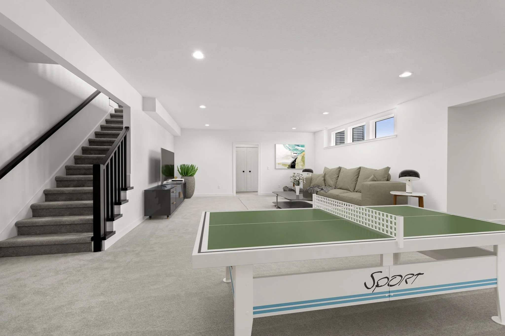 Finished basement recreation room with table tennis and seating area, carpeted floor, white walls, window lights, TV, potted plants, staircase, art, and lamps.