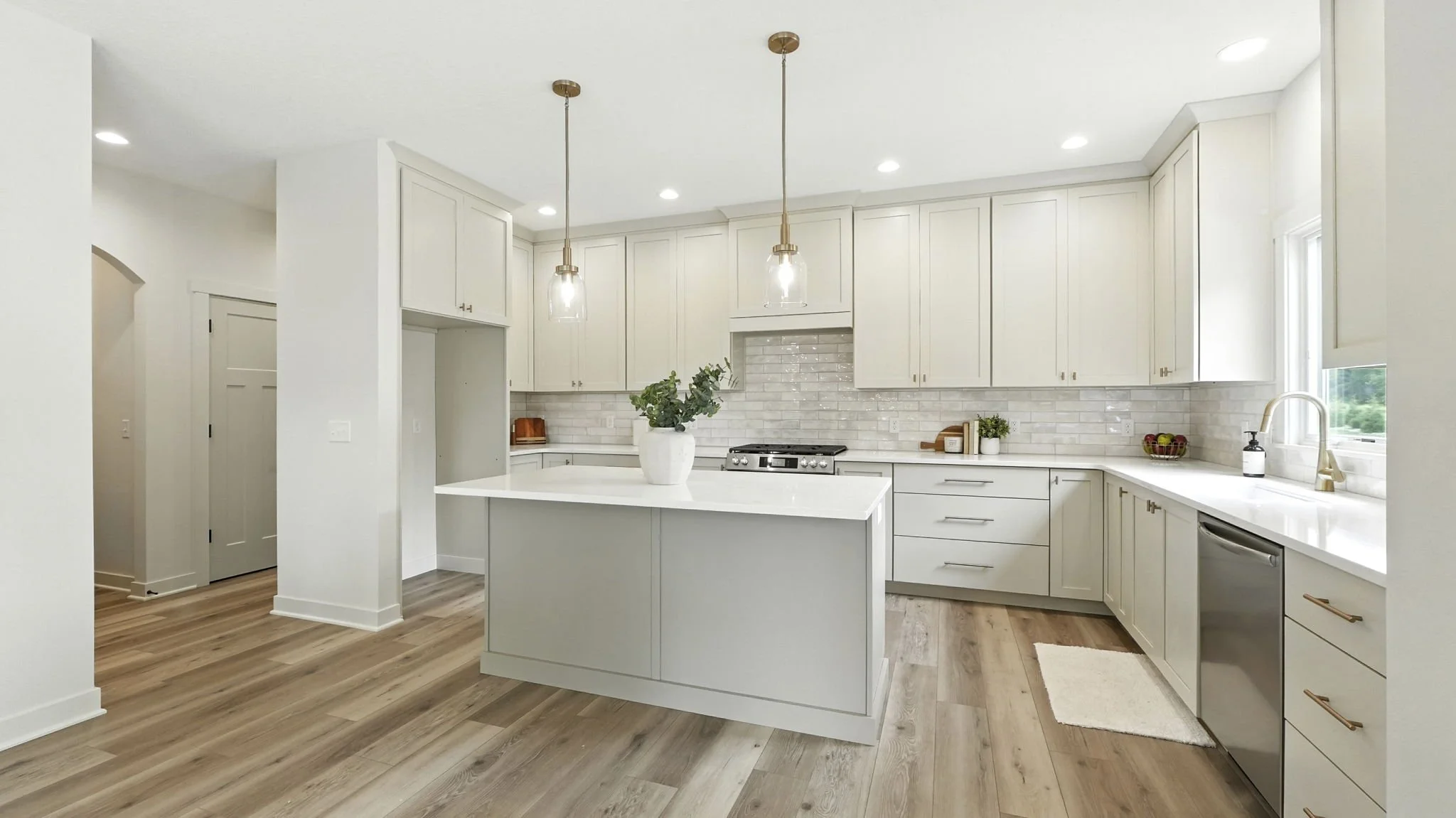 Modern white kitchen with island, pendant lights, wooden flooring, and natural light from window.
