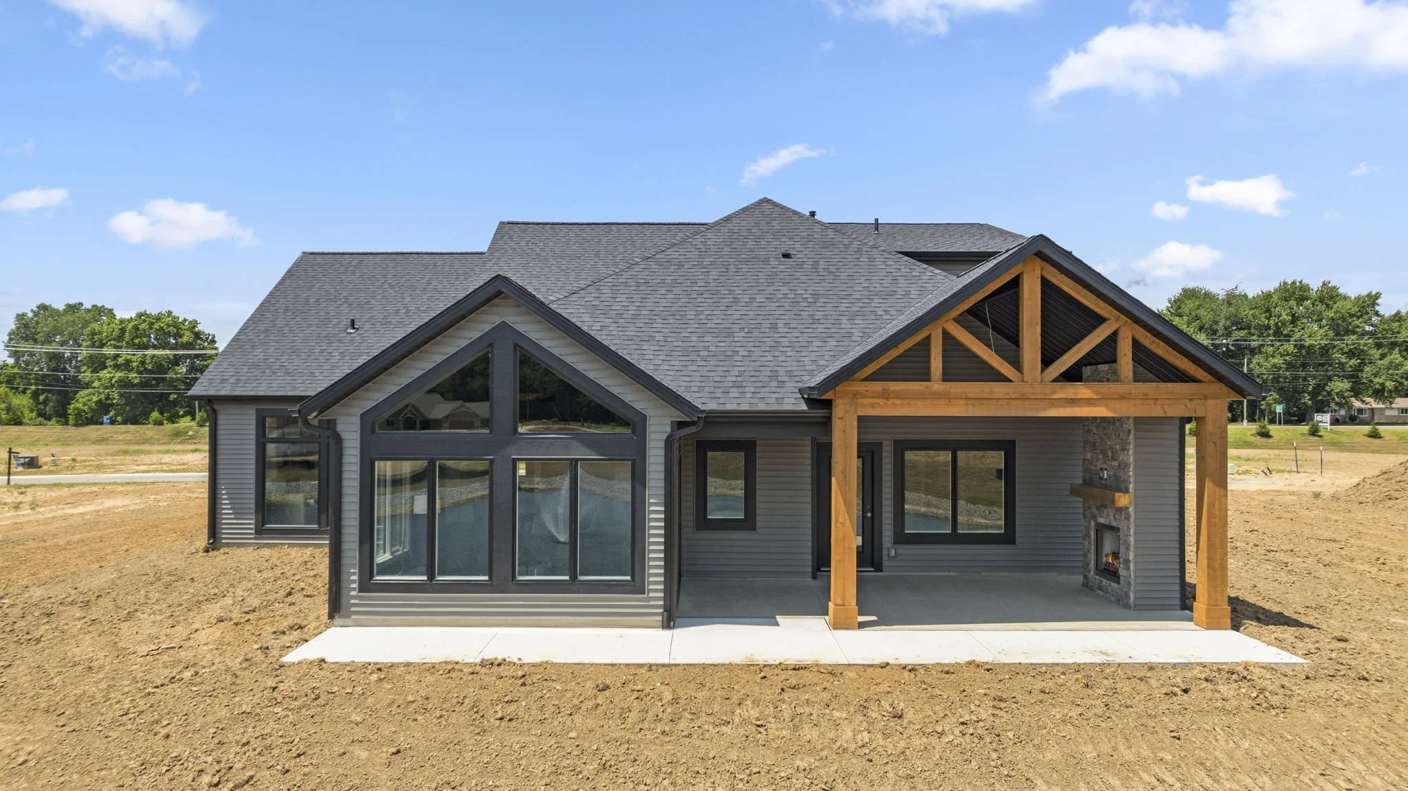 Modern house under construction with a gray exterior, large windows, and a covered patio with a stone fireplace. The house is built on a dirt lot with a blue sky and green trees in the background.