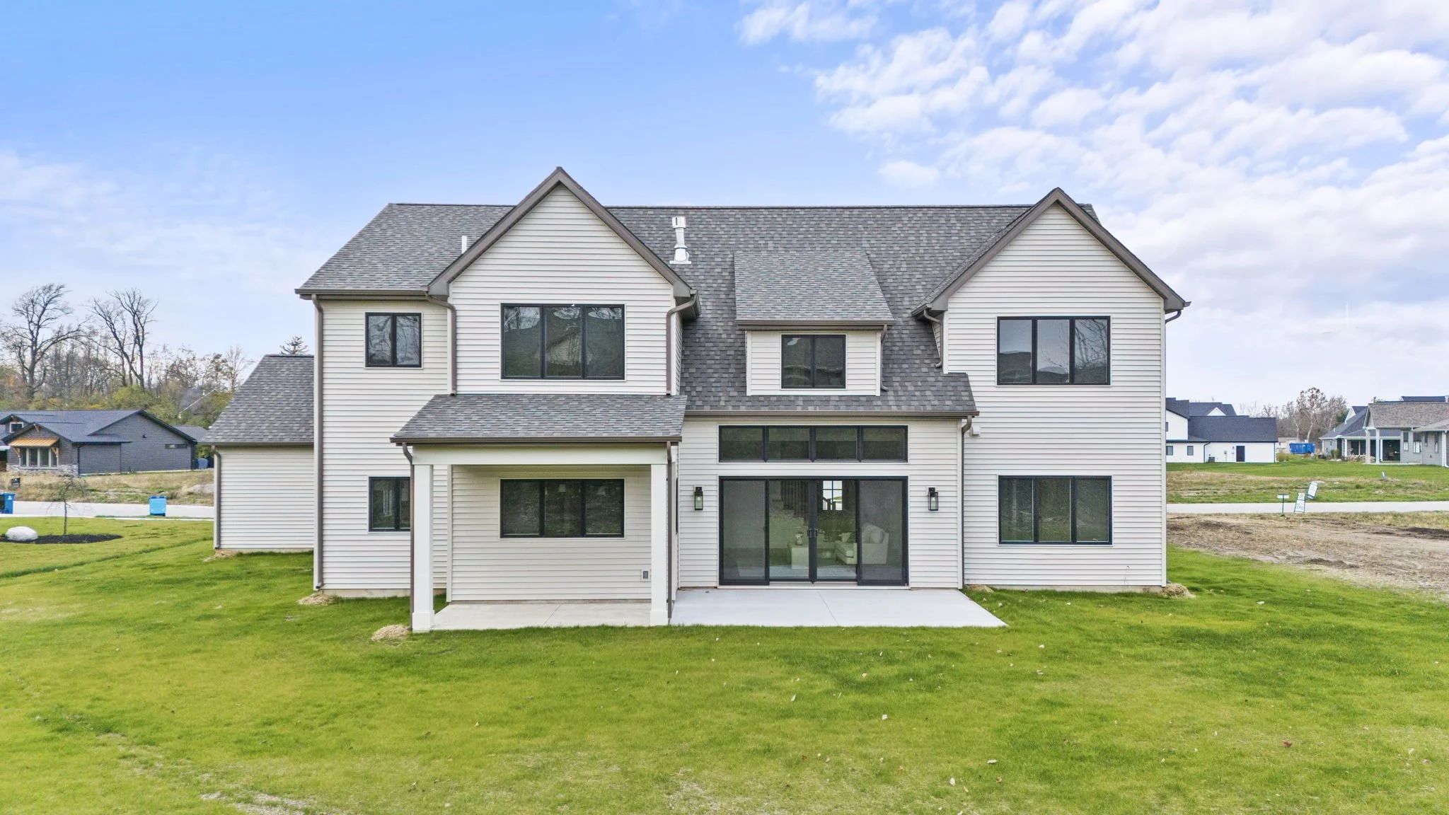 Rear view of a modern, two-story house with white horizontal siding, large windows, a sliding glass door, and a backyard with green lawn, under a partly cloudy sky.