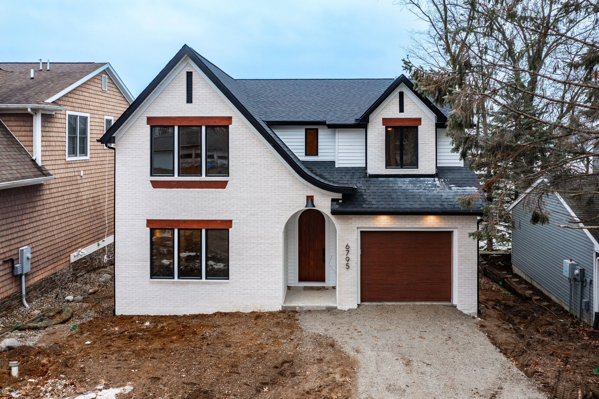 Newly constructed two-story house with white brick exterior, large windows, a wooden arched front door, and a one-car garage with a wooden door, surrounded by neighboring houses and leafless trees.