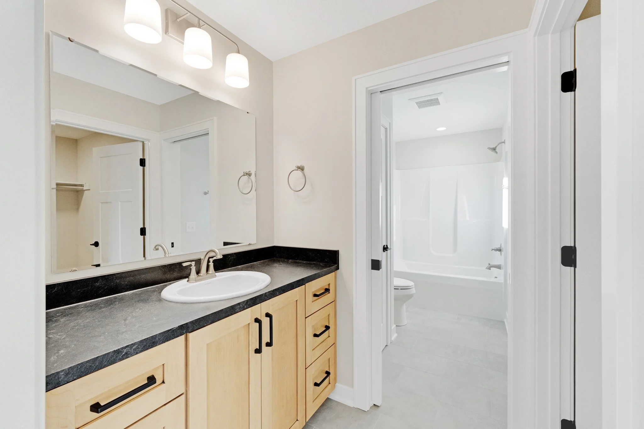 Bathroom with wooden vanity, black countertop, oval sink, large mirror, and modern light fixture; door to toilet and bathtub area.