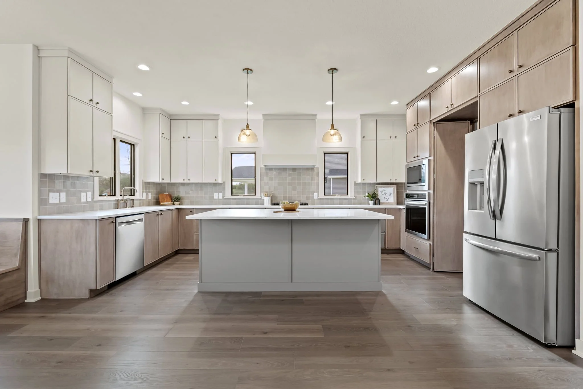 Modern kitchen with white and wood cabinets, stainless steel refrigerator, microwave, oven, and island with pendant lights, large windows, and wooden flooring.