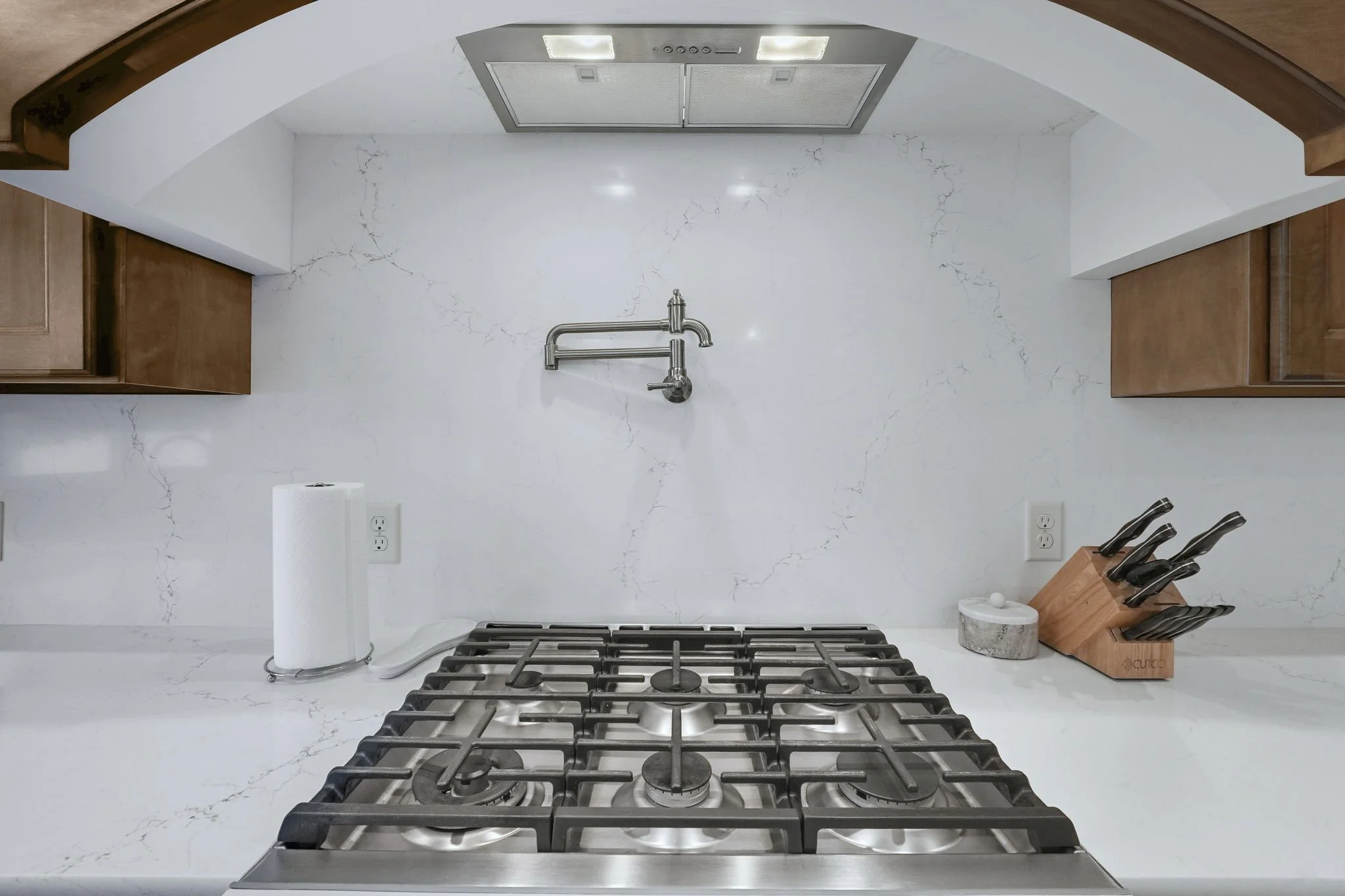 Kitchen with white marble countertop and backsplash, stainless steel gas cooktop, wooden cabinets, paper towel roll, and knife set in a wooden block.