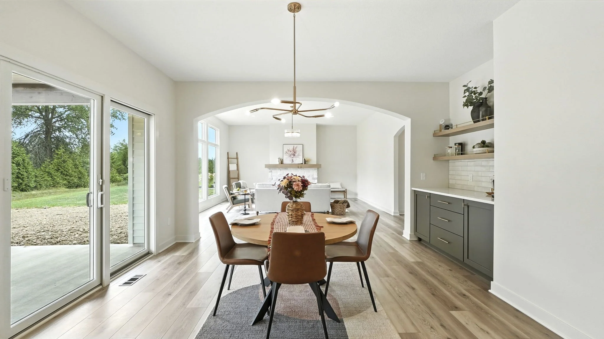 A dining area with a round wooden table and six brown chairs, a bouquet of flowers in a basket, and a gray and beige rug, leading to a bright living room with large windows, a white sofa, and a fireplace.