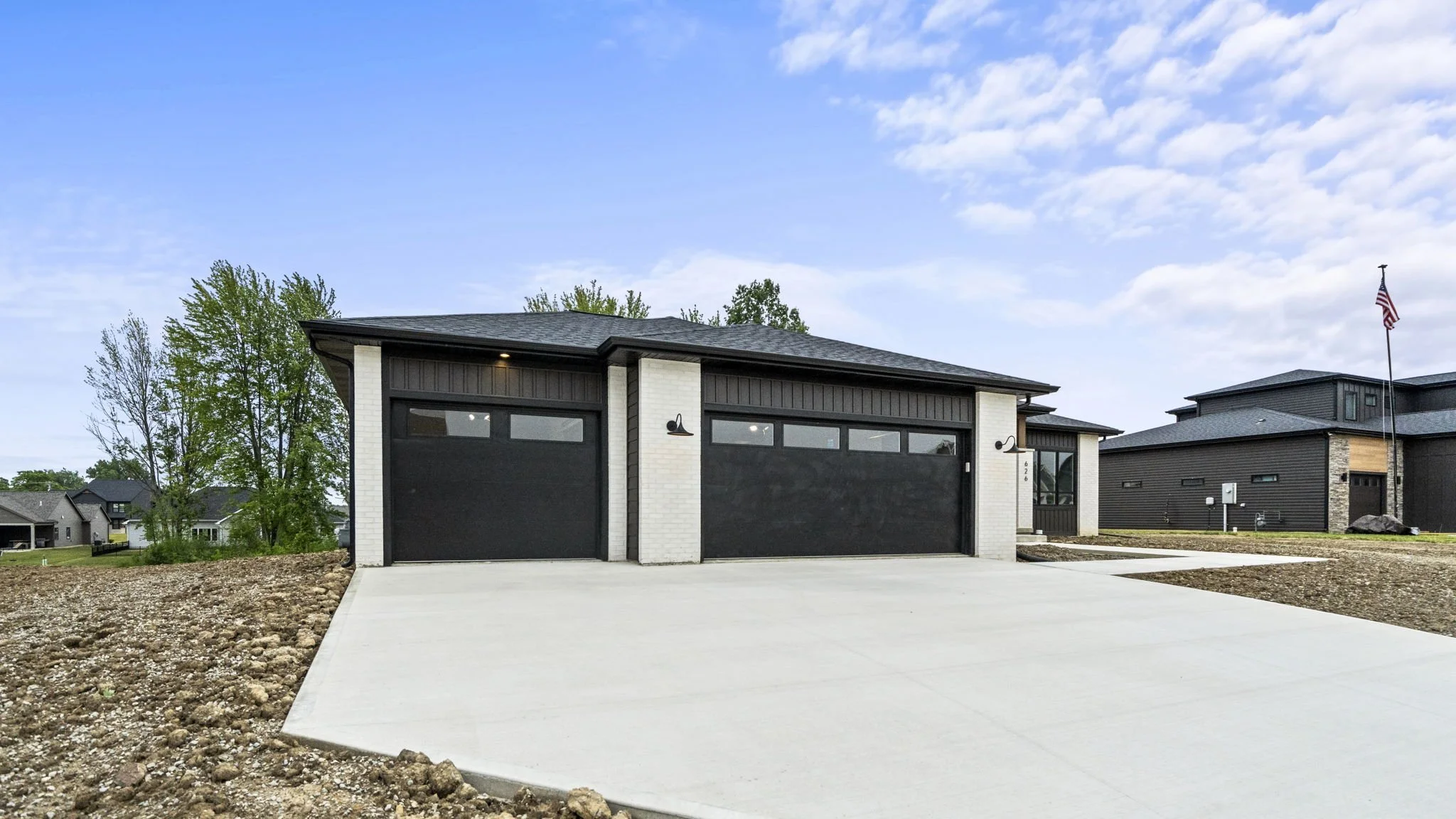 Modern house with black garage doors, white brick and black siding, concrete driveway, and a lawn with trees in the background, under a partly cloudy sky.
