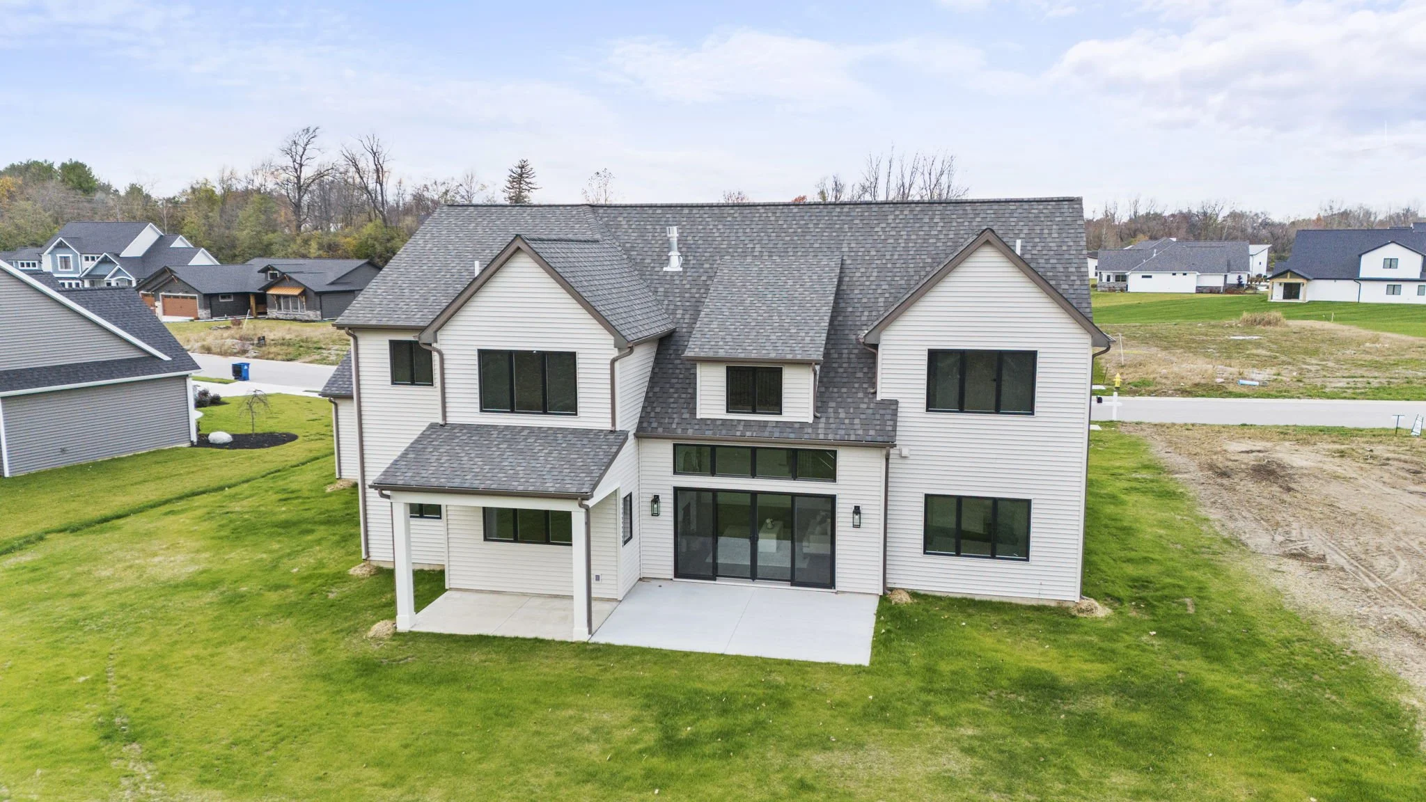 A modern white house with a large backyard and a concrete patio, surrounded by other houses and a green lawn, under a partly cloudy sky.