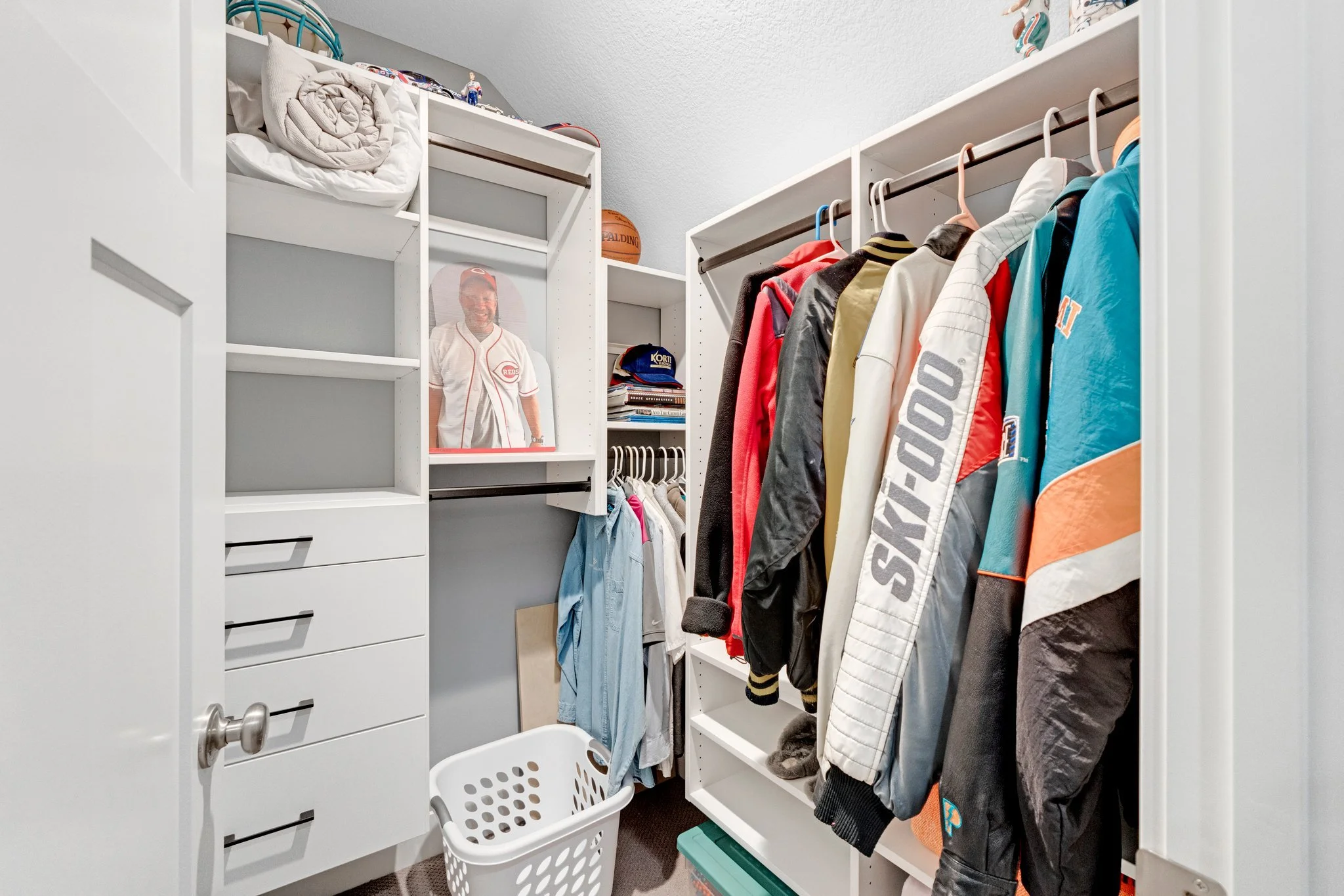 A walk-in closet with hanging jackets and shirts, a white laundry basket, and shelves holding miscellaneous items, including a framed photograph, baseball caps, and folded clothes.
