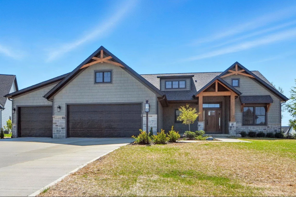 A modern two-story house with a gray exterior, stone accents, and a dark wood front door, featuring a three-car garage and landscaped front yard under a blue sky.