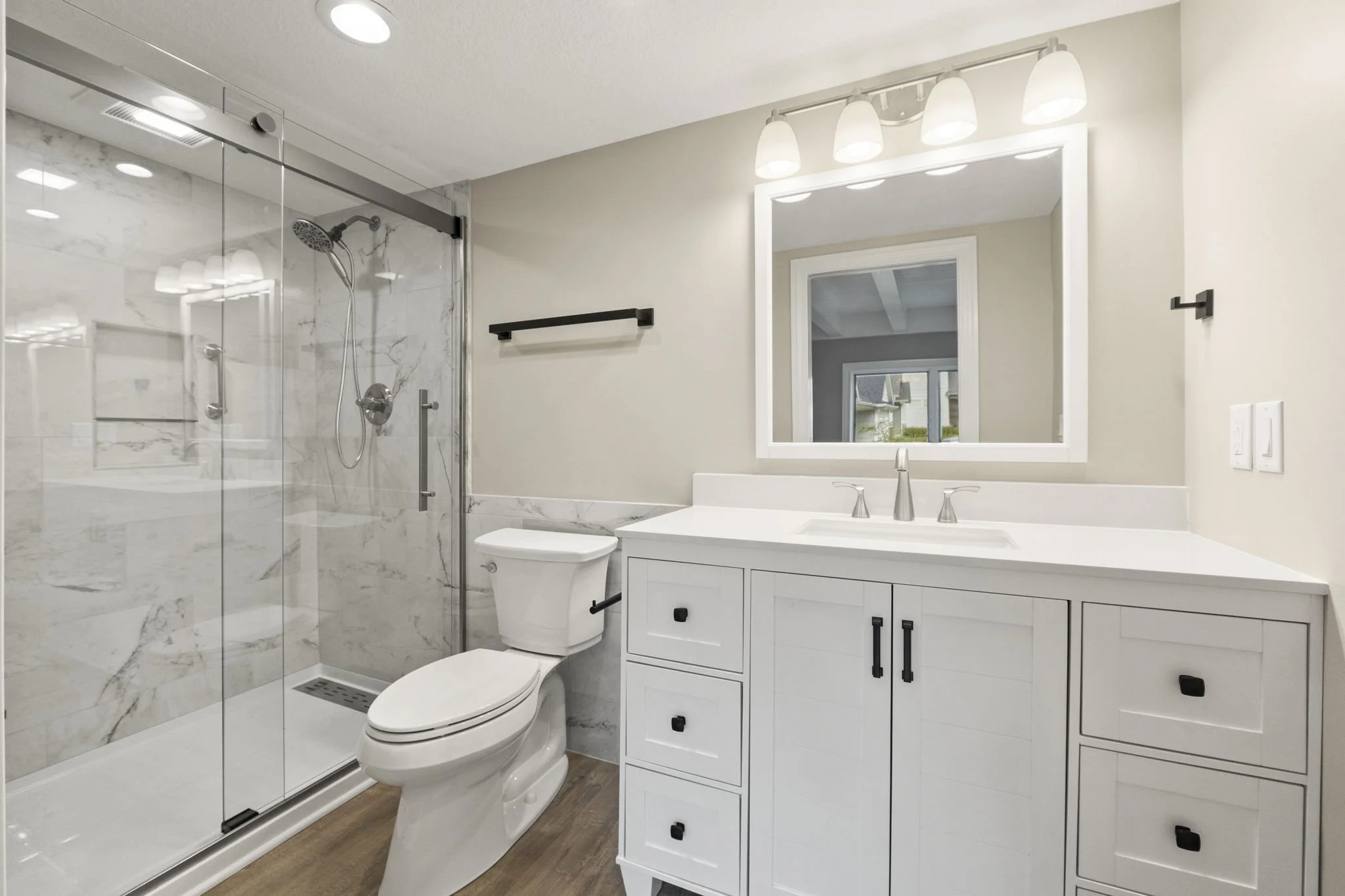 Modern bathroom with a glass-enclosed shower, white vanity with black handles, large mirror with built-in lighting, and a toilet. The shower has a rain shower head and marble wall tiles. The room has wood-like flooring and neutral wall colors.