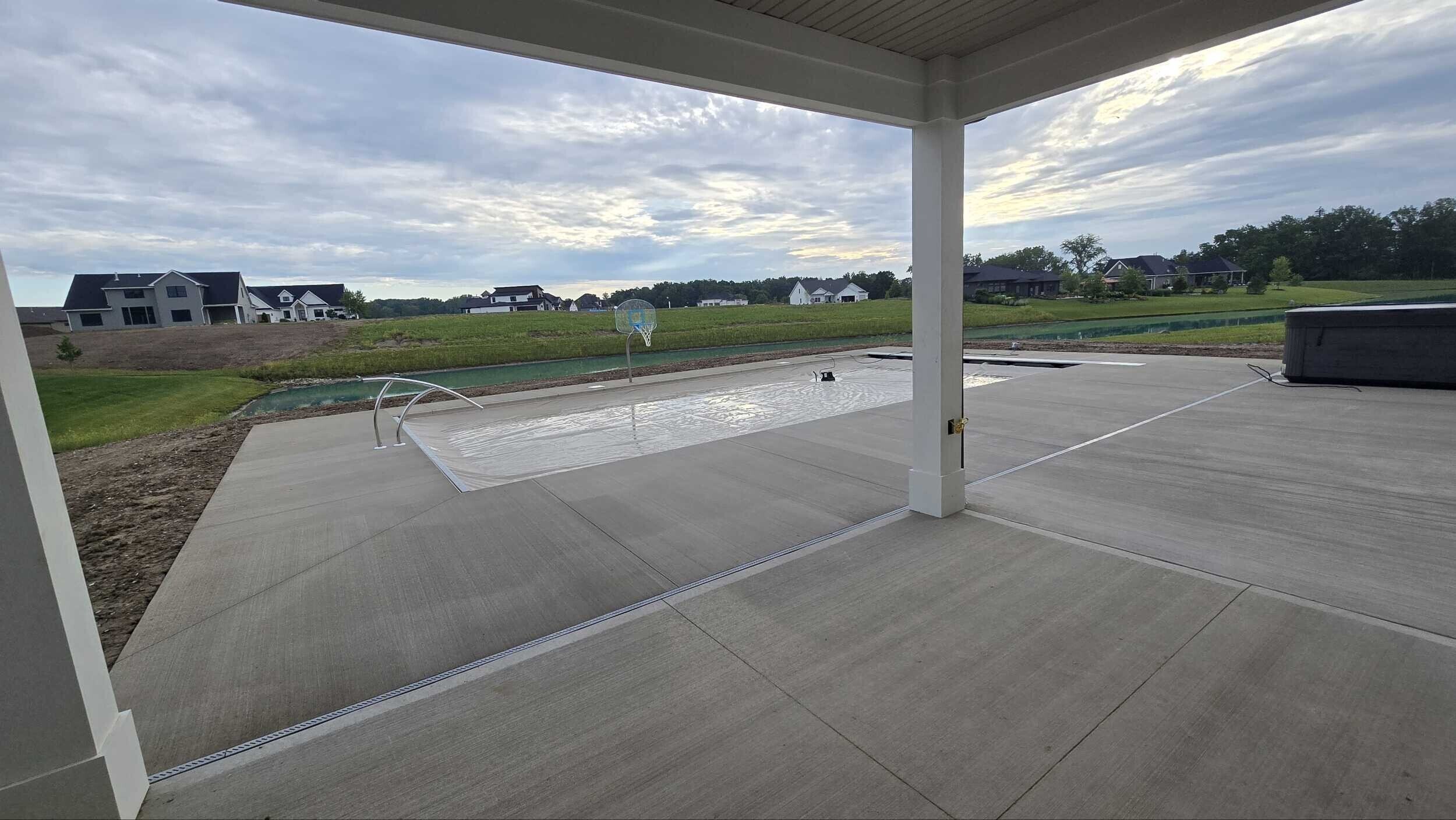 Backyard with swimming pool covered by a safety cover, with a basketball hoop and neighboring houses in the background under a partly cloudy sky.