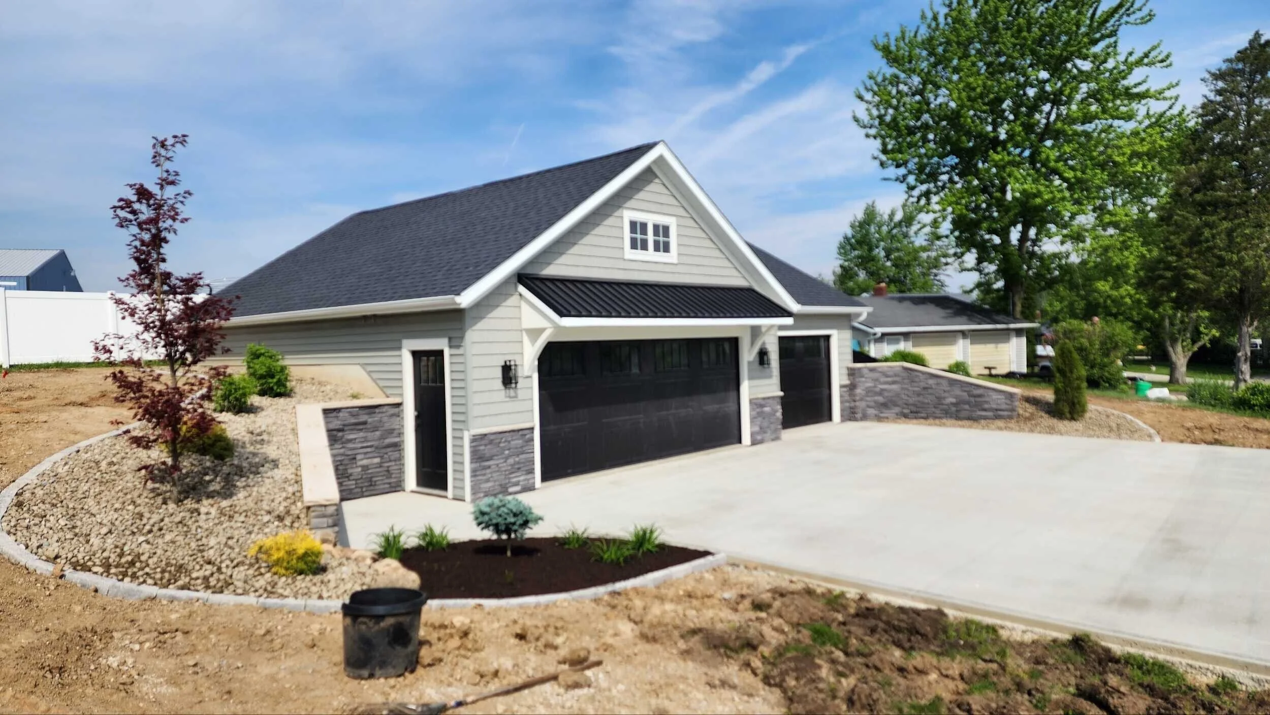 Newly constructed garage with a black door, gray siding, stone accents, and surrounding landscaping with trees and shrubs, in a suburban neighborhood.