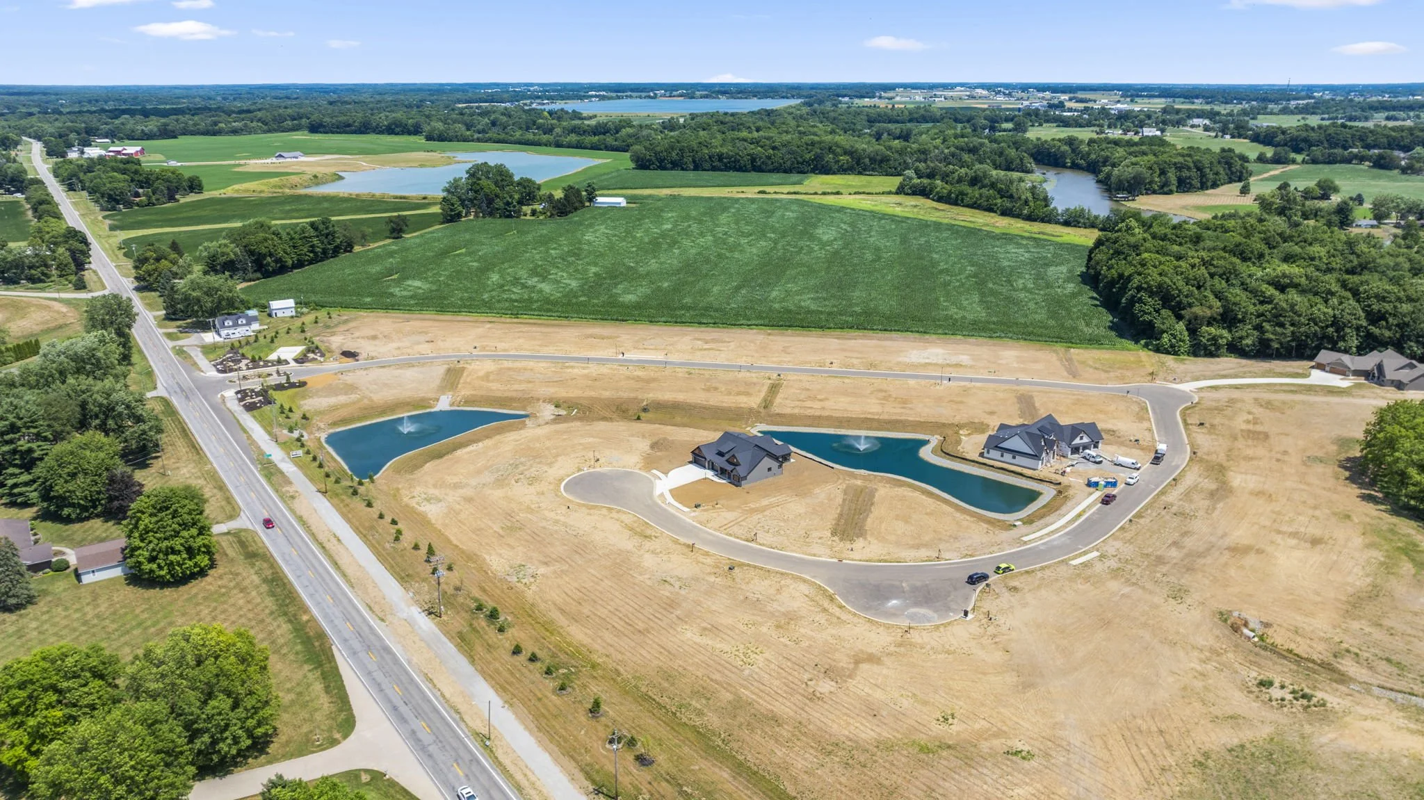 Aerial view of a developing residential area with a curved road, water features, and houses under construction amid open land and green fields.