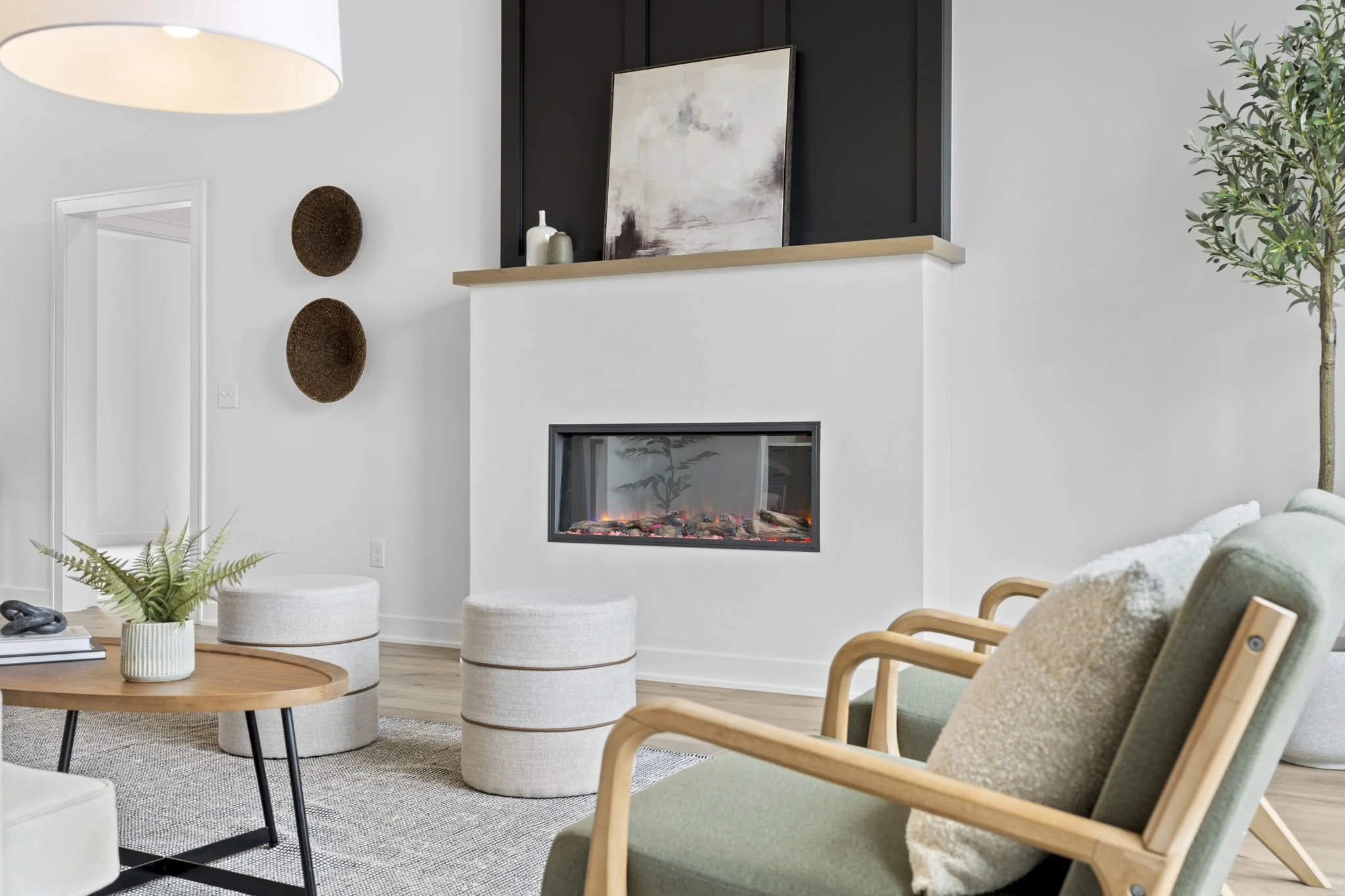 Modern living room with green armchair, light-colored pillows, wooden coffee table with a plant, two beige ottomans, a white fireplace, black wall above fireplace with artwork and vases, decorative woven wall pieces, and a tall plant in the corner.