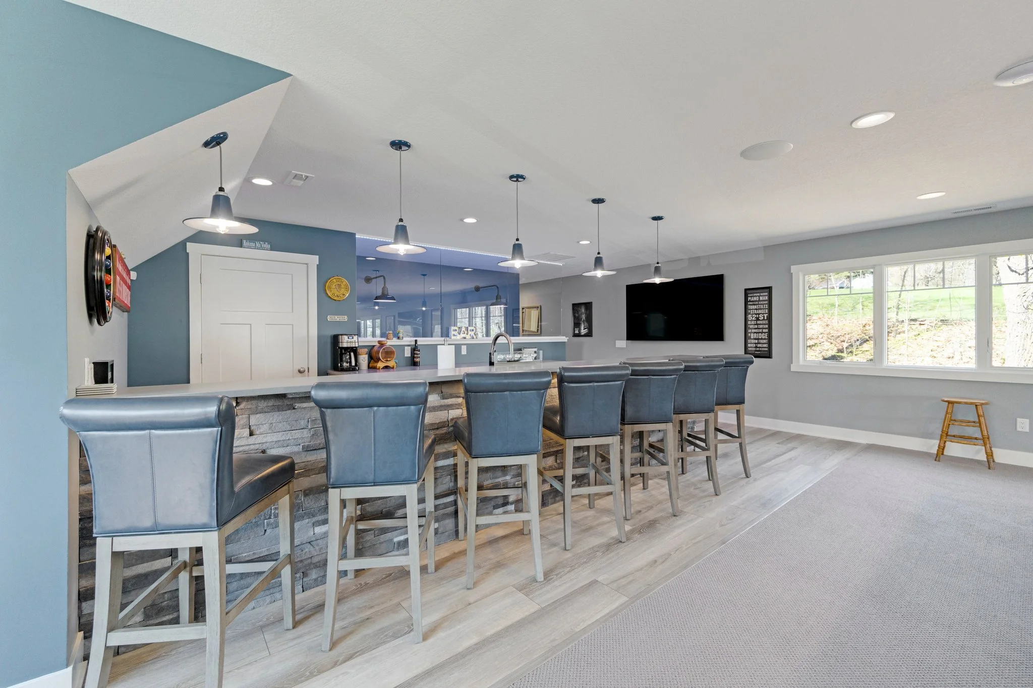 Modern basement bar area with a stone front counter, six gray bar stools, pendant lights, a large TV, a window, and a small wooden stool.