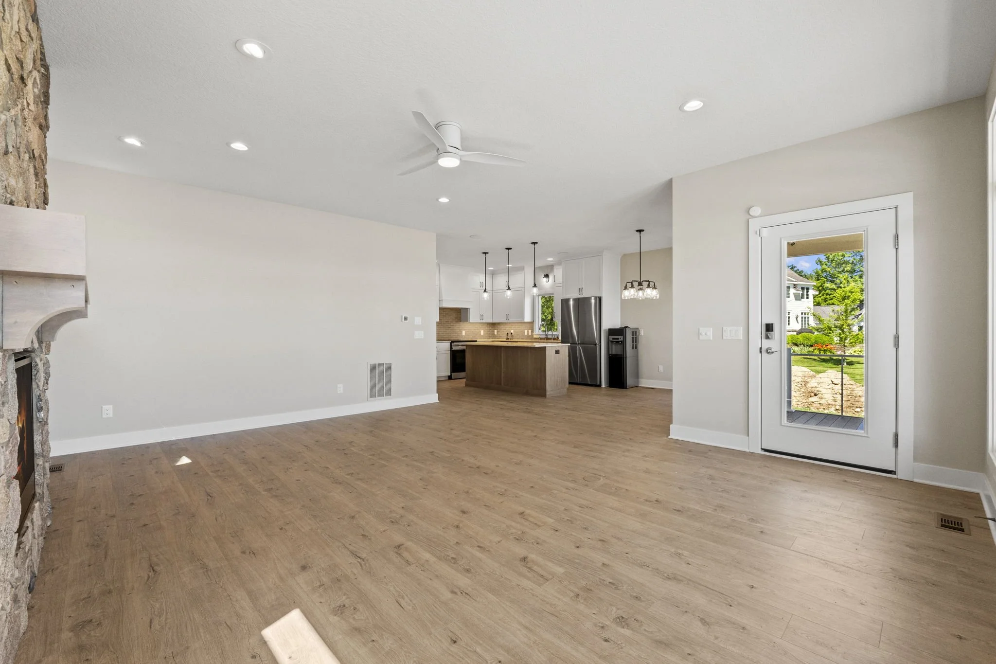 Open concept living space with wood floors, kitchen area with island, stainless steel refrigerator, and a glass door leading outside to a yard with green trees and bushes.