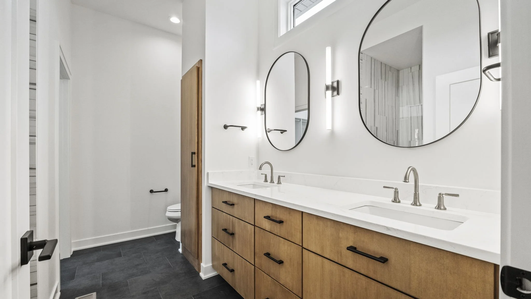 Modern bathroom with double vanity, wooden cabinets, white marble countertop, two oval mirrors, and black hardware; in the background, a separate toilet area with white walls, a small window, and a black towel bar.