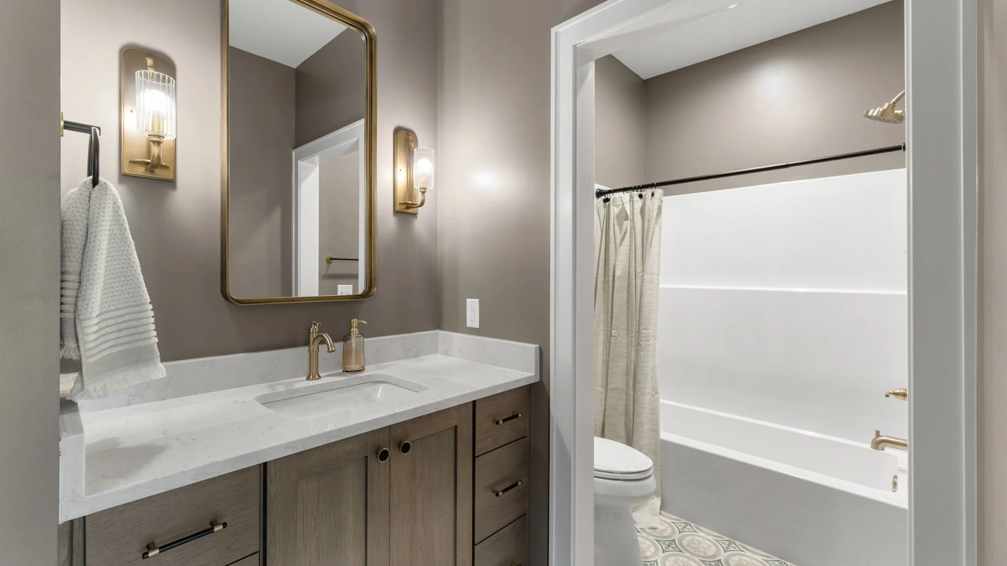Bathroom featuring a vanity with a marble countertop, a wooden cabinet with gold accents, a rectangular mirror with a gold frame, wall sconces with gold fixtures, a towel hanging on a black rod, and a shower/tub area separated by a partial wall, with