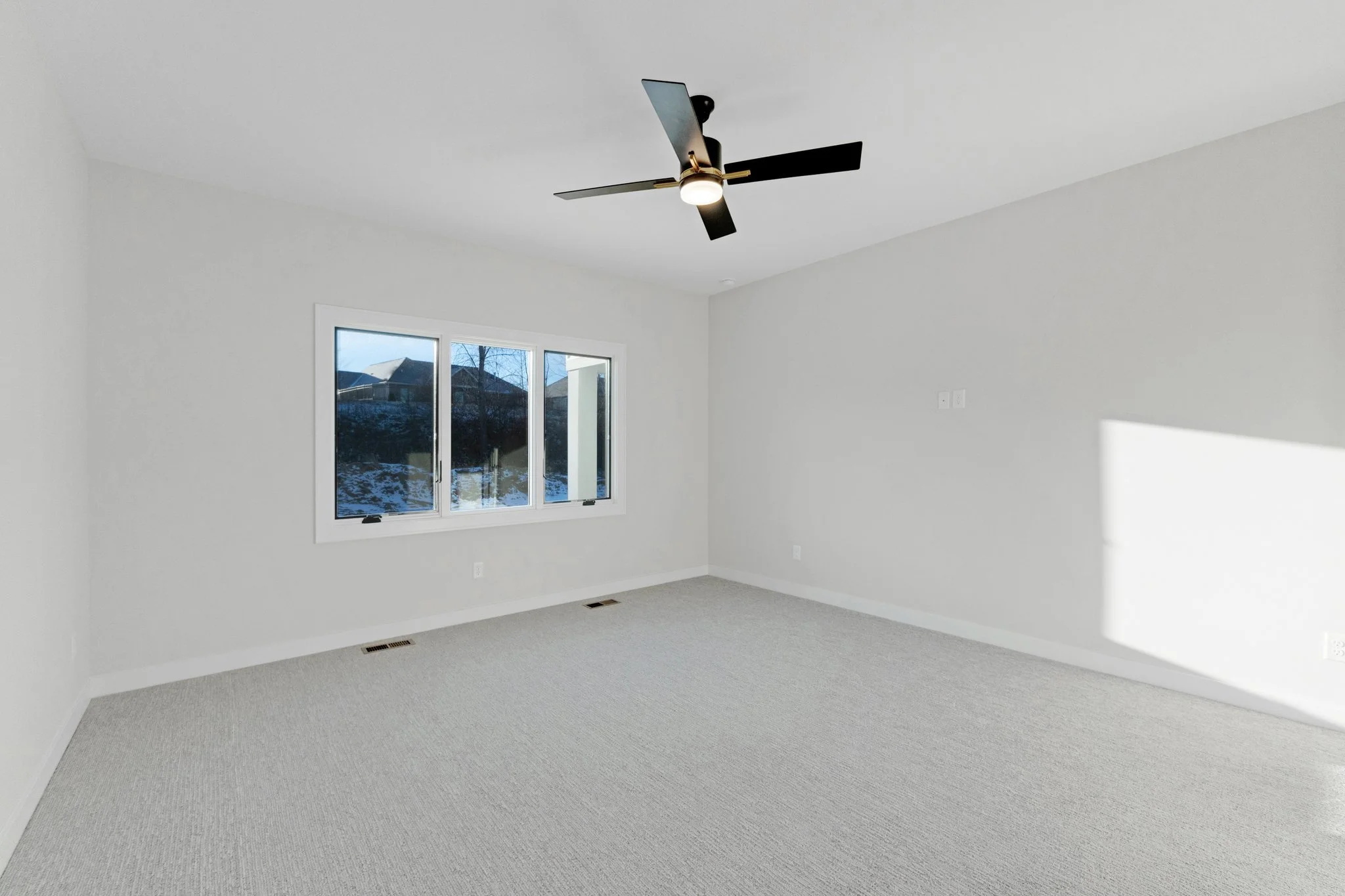 Empty room with white walls, beige carpet, a window showing a snowy landscape outside, a black ceiling fan with a light, and sunlight on the right wall.