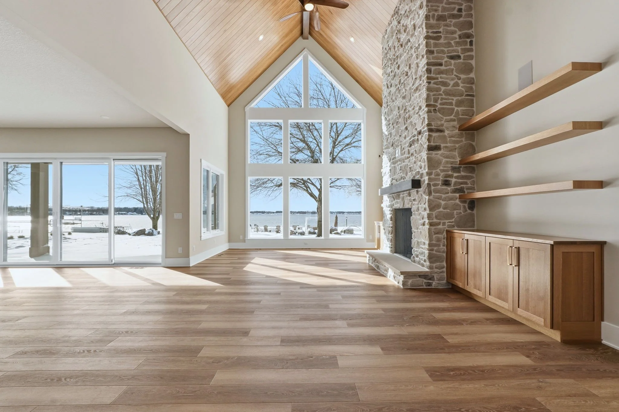 Living room with large front windows overlooking snowy landscape, stone fireplace, wooden shelves, and hardwood flooring.