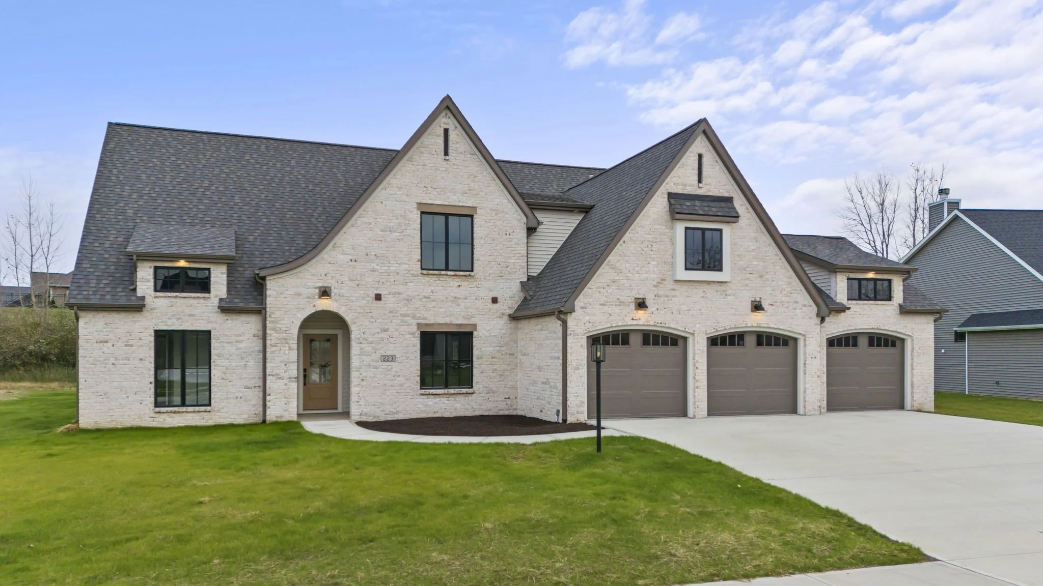 A modern two-story house with a brick exterior, arched doorway, three-car garage, and a well-maintained lawn under a partly cloudy sky.