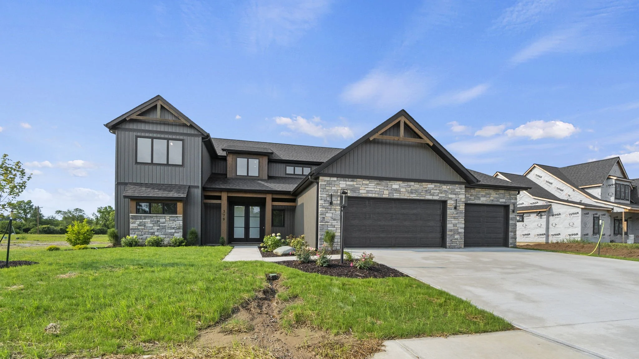 Modern two-story house with a mix of dark siding, stone accents, and large windows, under a blue sky with some clouds, with a green lawn and driveway in front.