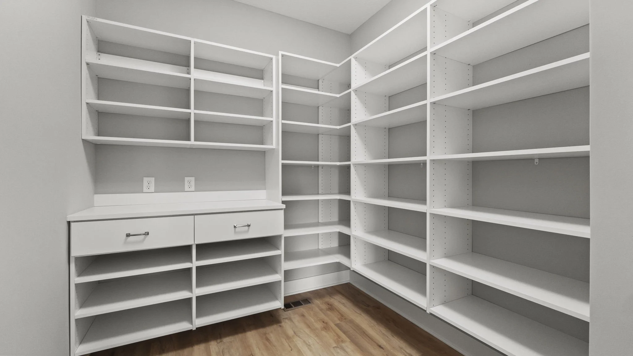 Empty white built-in pantry with multiple shelves and drawers, beige walls, hardwood flooring, and wall outlets.
