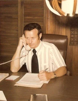 A man in a short-sleeved, striped shirt and black tie sitting at a desk, talking on a corded telephone, holding a pen over papers.