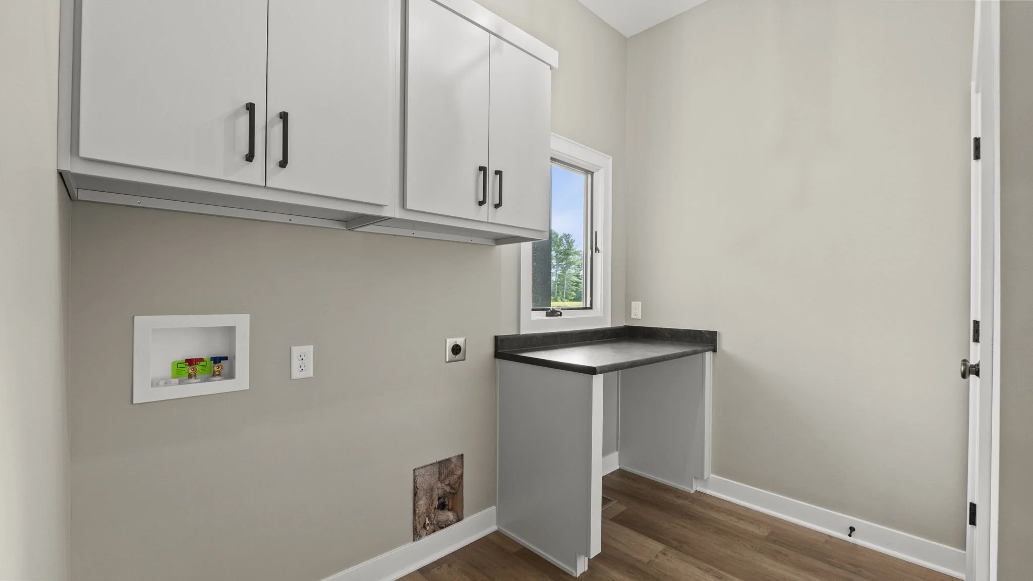 Empty laundry room with white cabinetry, a black countertop, a small window, and unfinished plumbing connections on the wall.