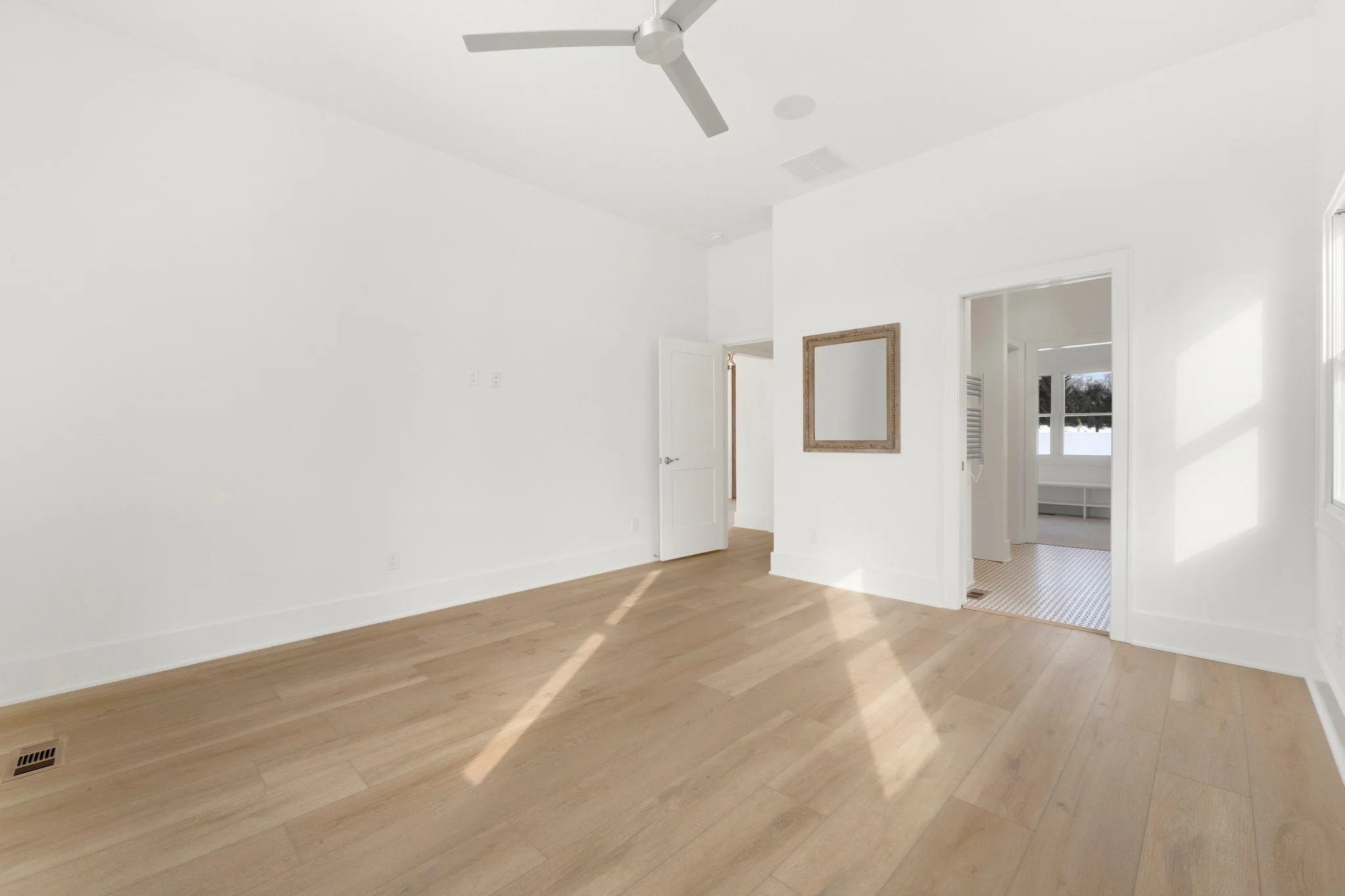 Empty bright room with white walls, wooden floor, ceiling fan, and sunlight coming through windows.