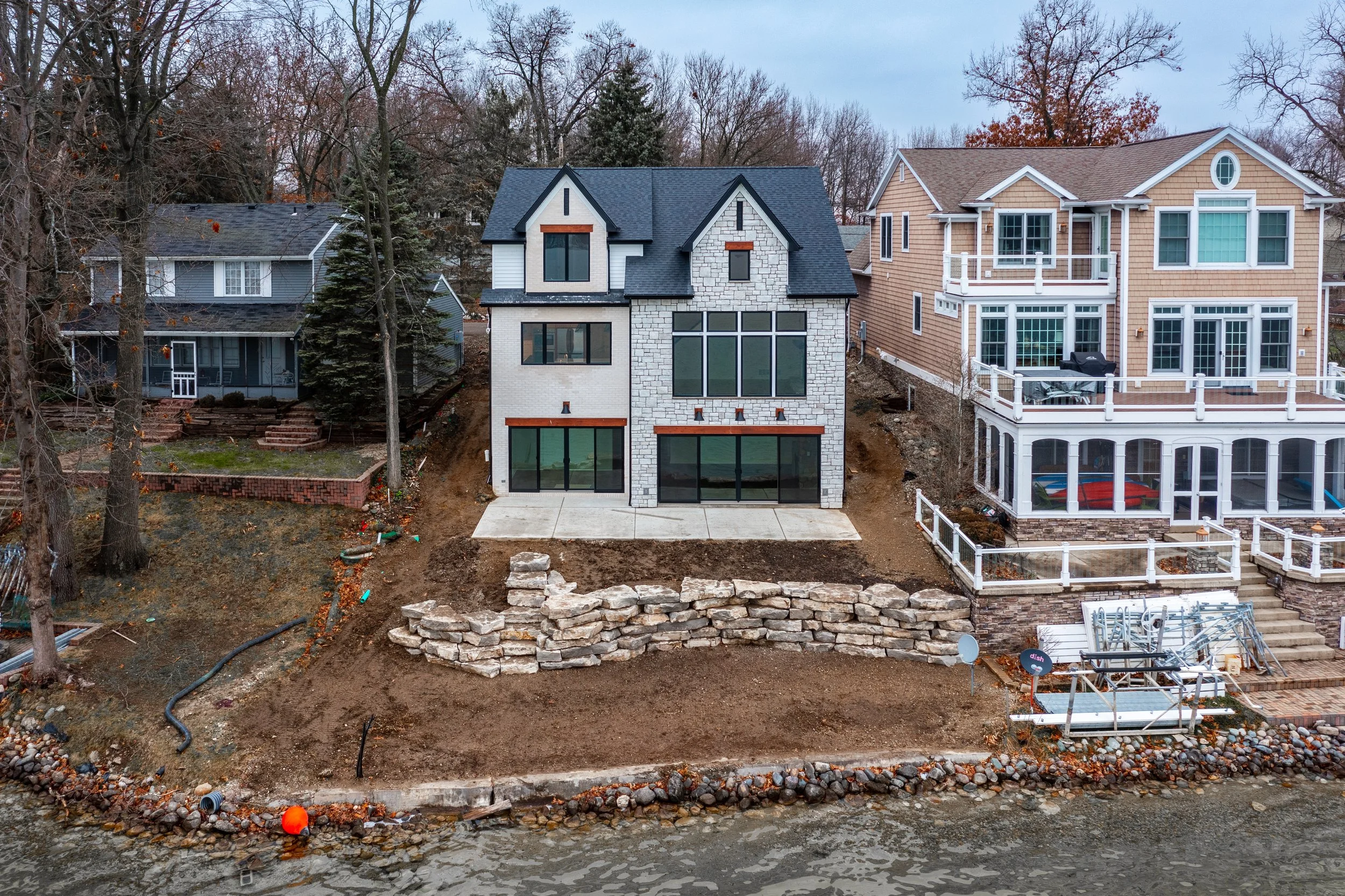 A newly constructed modern house on a waterfront lot with exposed soil and a stone retaining wall, flanked by neighboring houses on a leafless trees-lined street in late fall or winter.