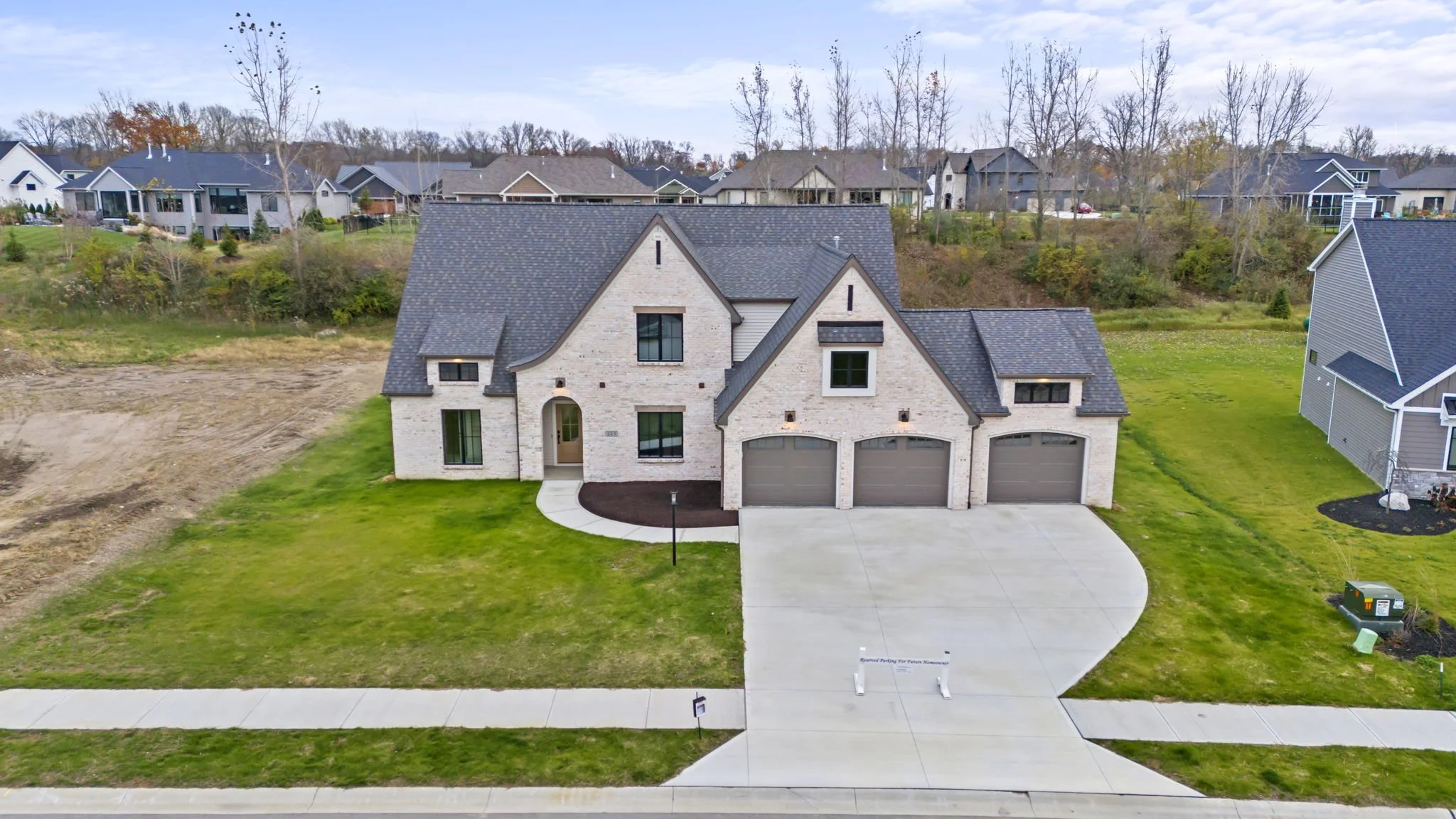 A newly constructed two-story house with white brick exterior, gray roof, three-car garage, and front lawn, situated in a suburban neighborhood with other houses in the background.