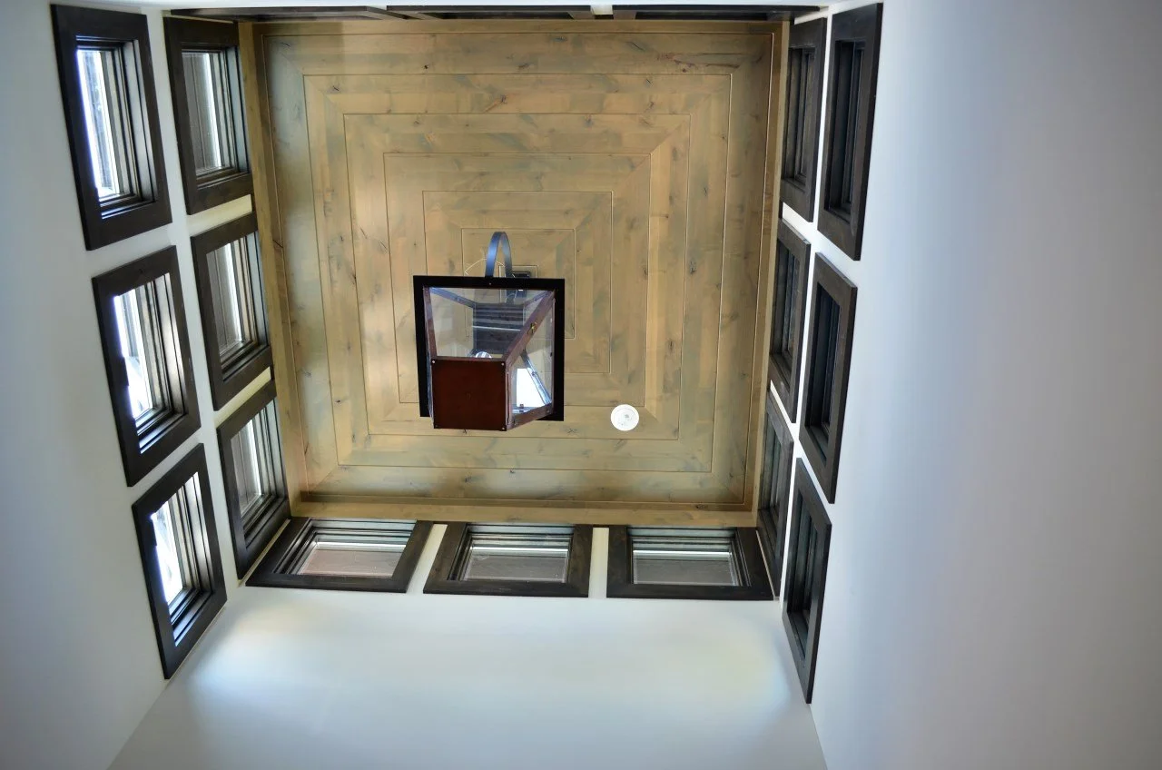 View of a ceiling with a hanging glass light fixture, surrounded by a wooden ceiling. Walls are white with multiple black-framed windows.