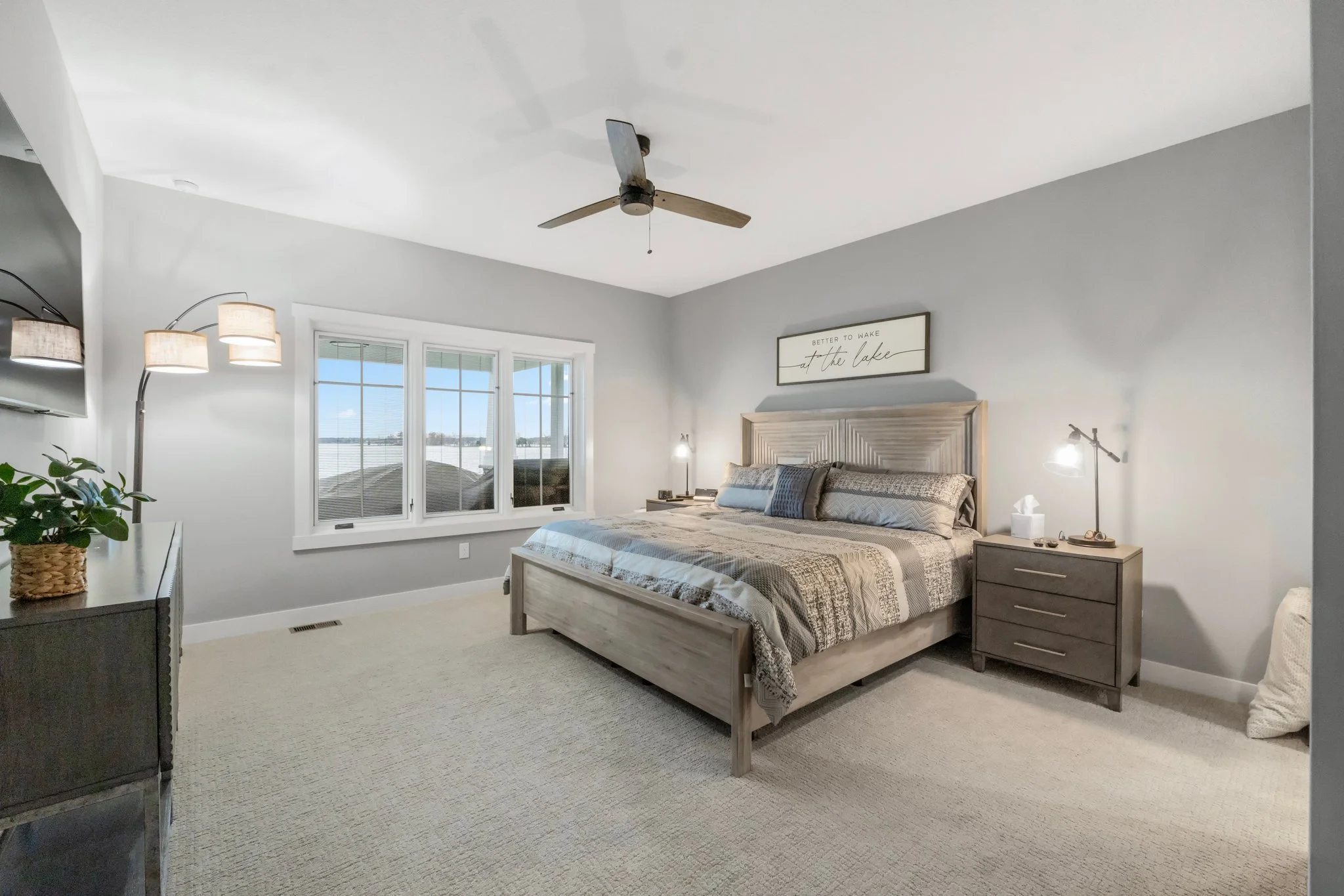 Bright bedroom with large window overlooking water, cozy bed with patterned bedding, dark nightstands with lamps, ceiling fan, and simple wall decor