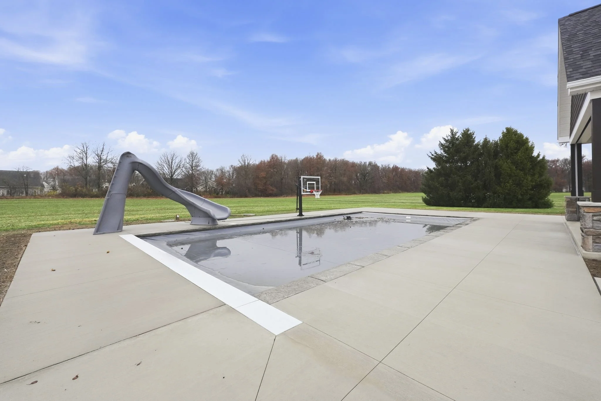 An empty swimming pool with a slide and basketball hoop in a backyard on a partly cloudy day.