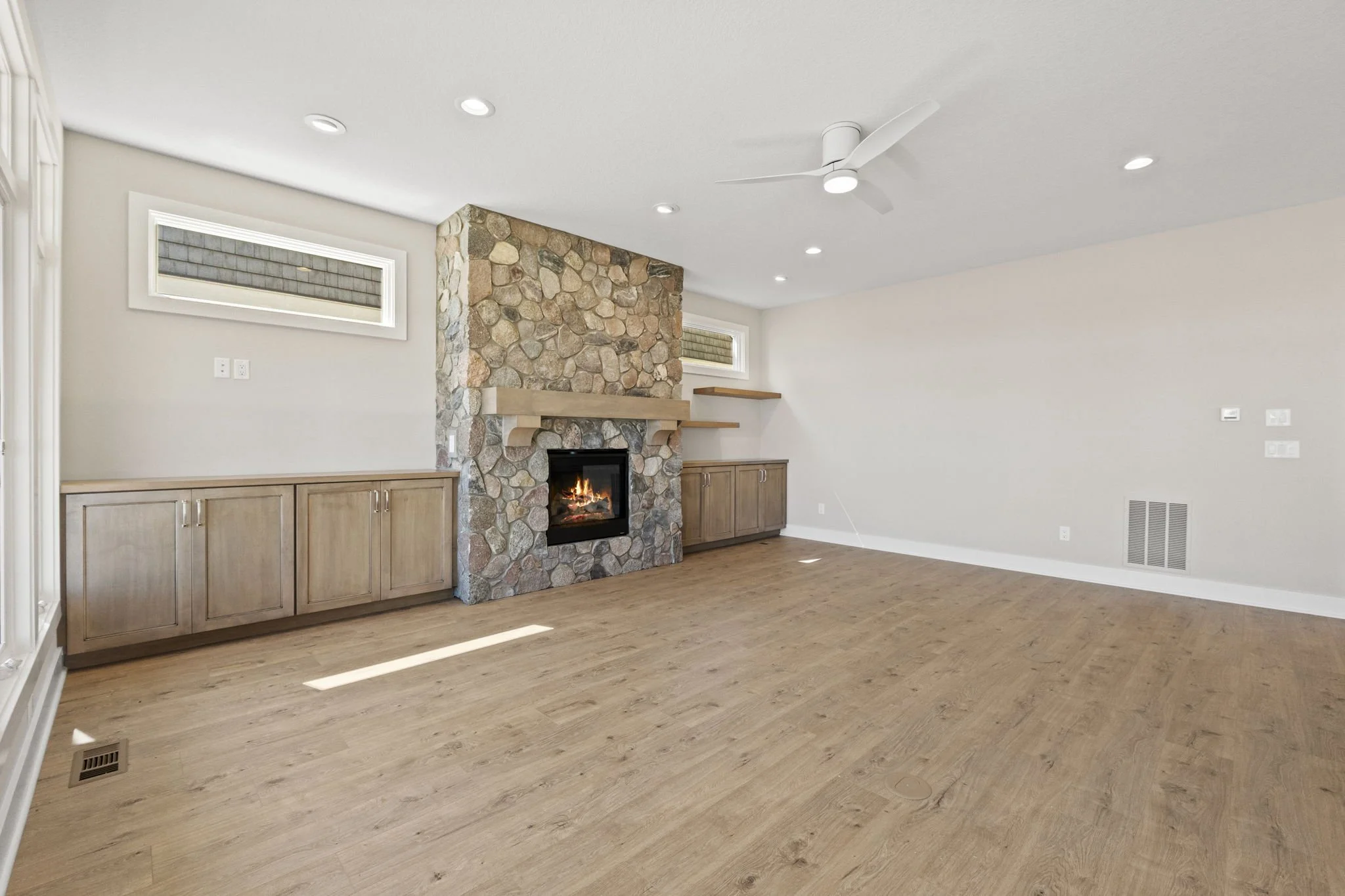 Empty living room with a stone fireplace, wooden built-in cabinets, hardwood floors, ceiling fan, white walls, and small high windows.