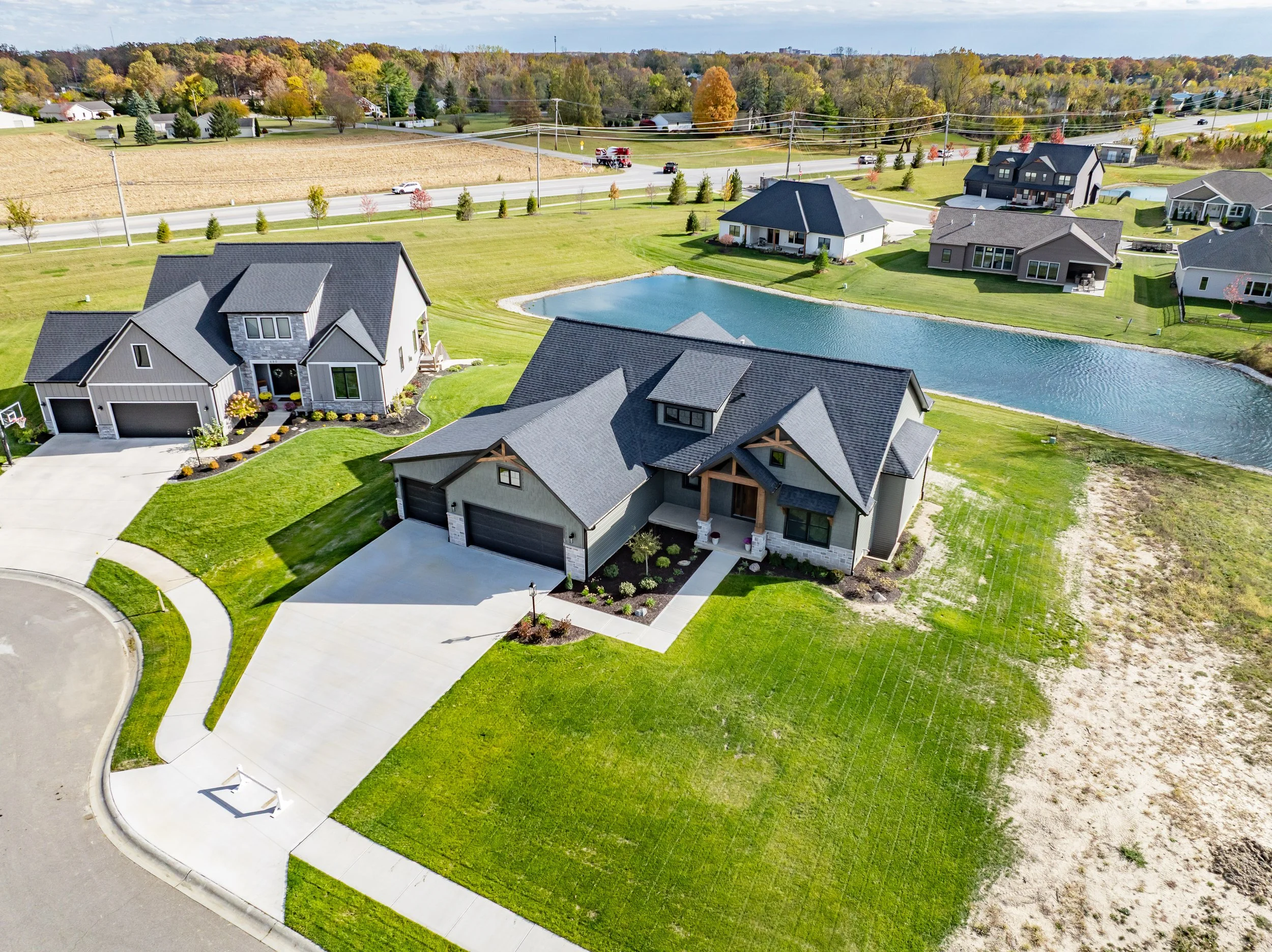 Aerial view of a residential neighborhood with modern houses, green lawns, a pond, and trees in the background.