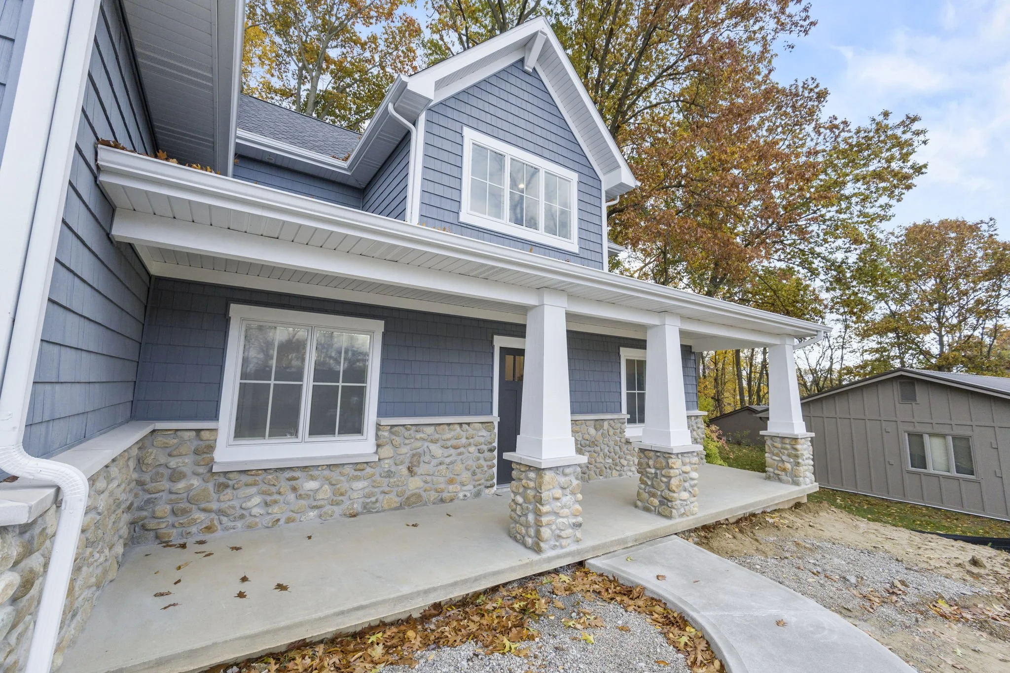 A newly built two-story house with blue siding, stone accents on the lower walls, and a covered front porch with white columns. The house is surrounded by trees with autumn foliage and a clear sky.