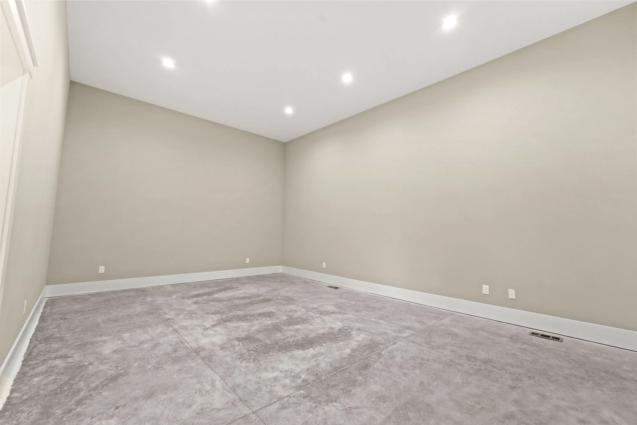 Empty beige-colored room with a concrete floor, ceiling lights, and electrical outlets along the walls.