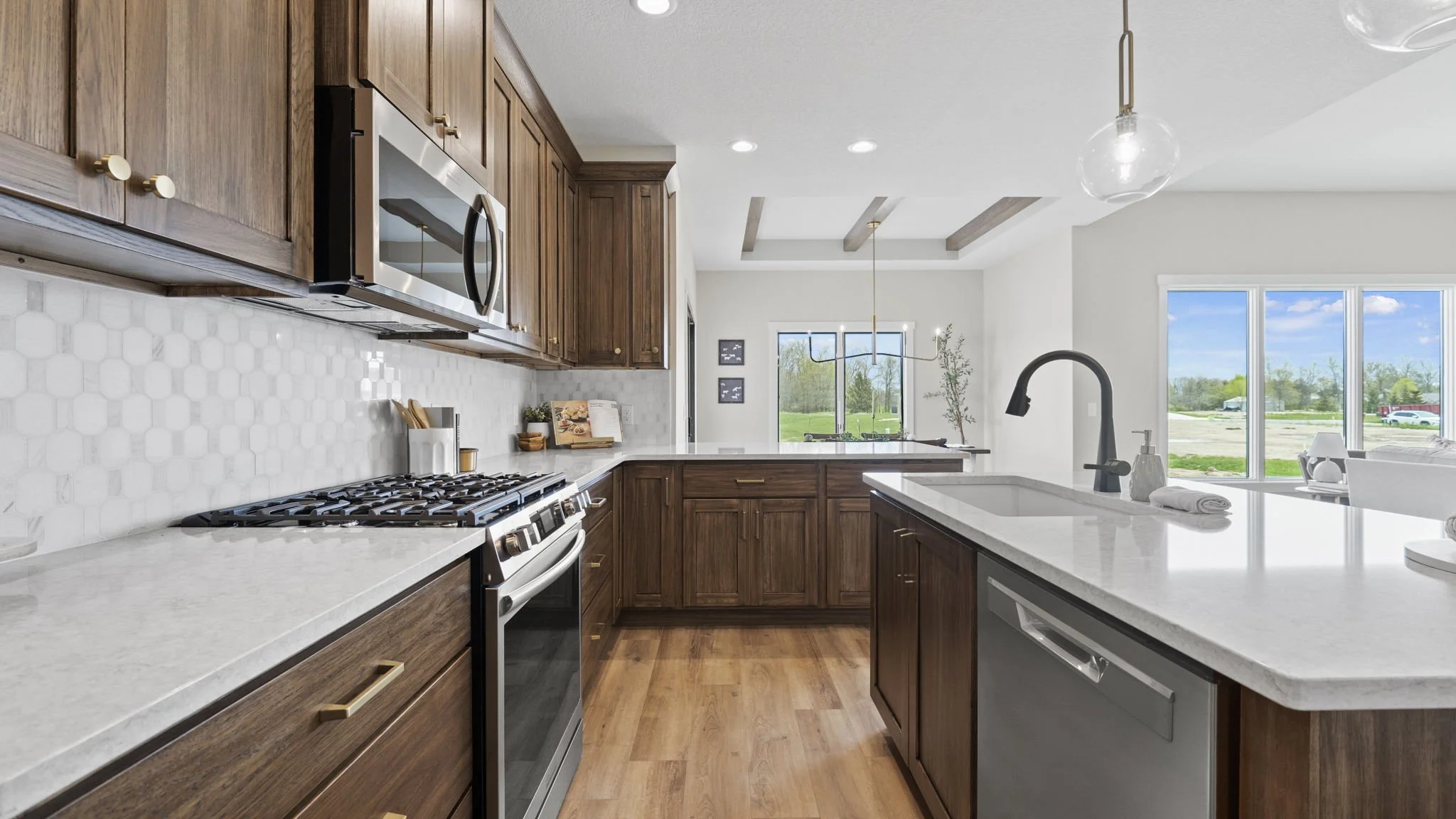 Modern kitchen with wooden cabinets, white quartz countertops, stainless steel appliances, and a view of a green outdoor landscape through large windows.