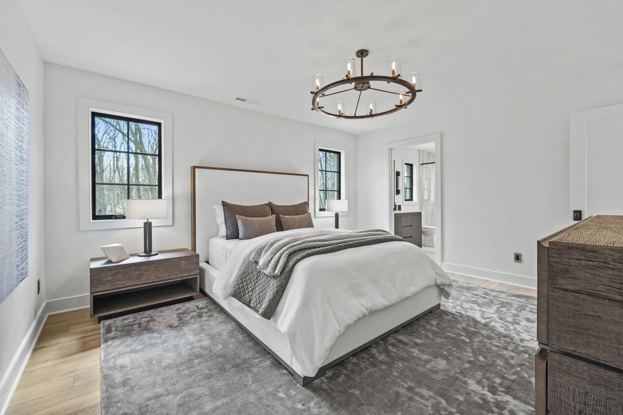 Bright bedroom with white walls, large bed with white linens, multiple brown and beige pillows, gray throw blanket, bedside tables with lamps, two windows with black frames showing outdoor trees, a chandelier style light fixture, a doorway leading to