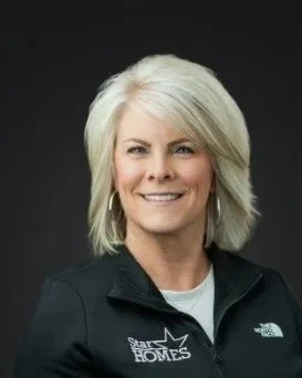 Portrait of a woman with blonde hair smiling, wearing a black North Face jacket with a 'Start With Homes' logo, against a dark background.