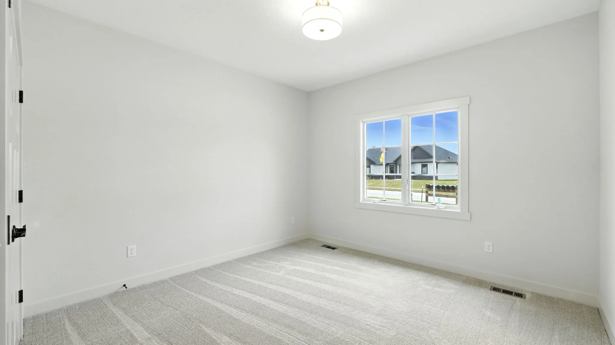 Empty room with white walls, beige carpet, large window showing outside houses, ceiling light fixture, and heater vents.