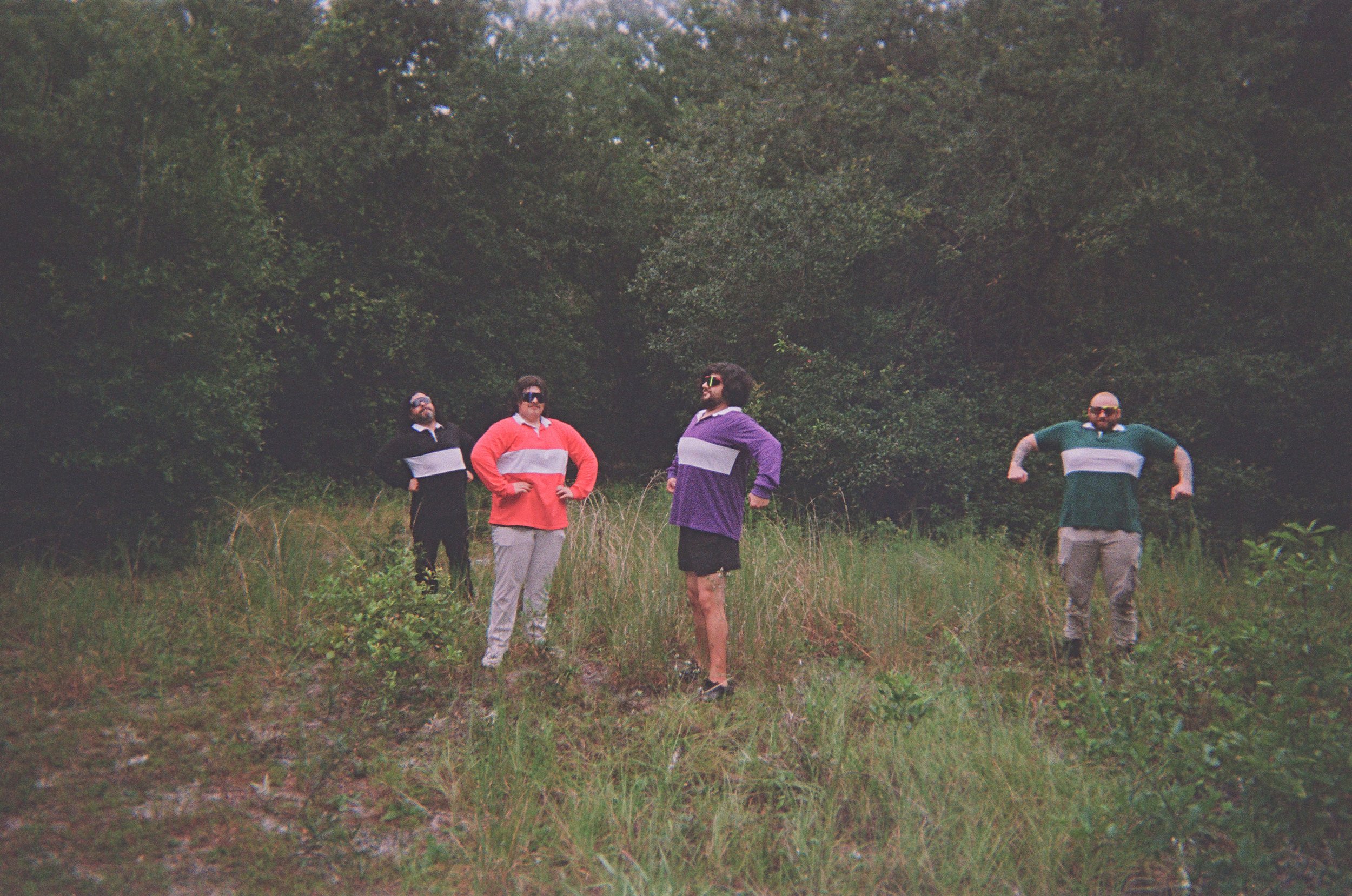 Four men dressed in colorful sweaters and casual pants or shorts, standing outdoors in a grassy area with trees in the background, striking poses and looking in different directions.
